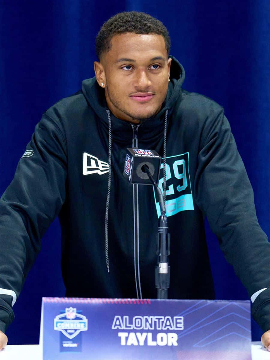  壁紙 — 一位身穿黑色連帽衫的年輕NFL球員坐在記者會桌前，桌上放有麥克風和名牌。