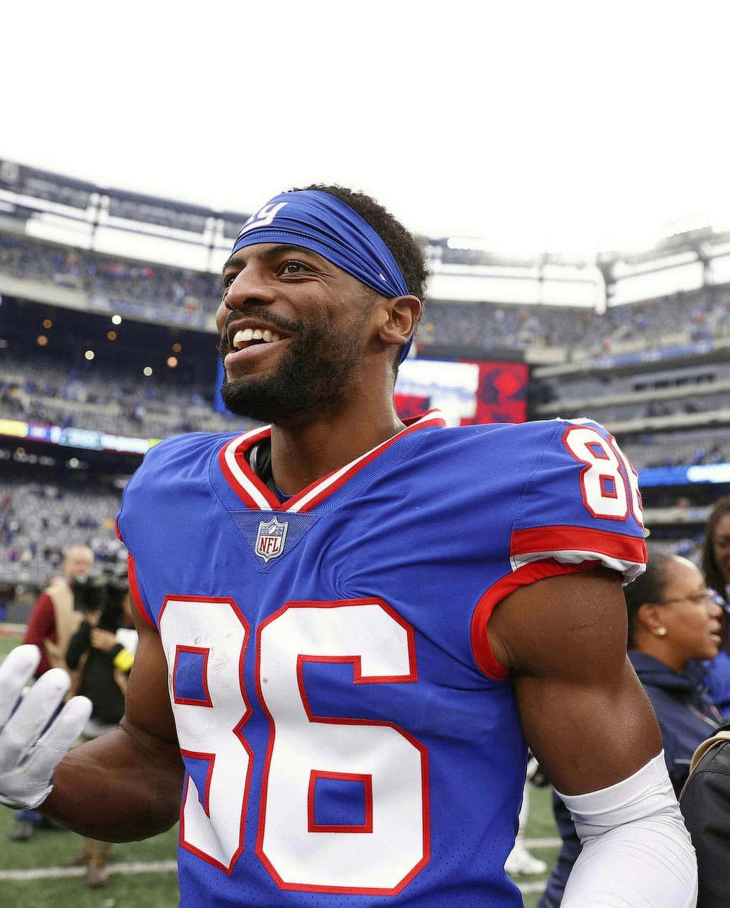  壁紙 — 一位身穿藍色球衣、號碼86的NFL球員面帶微笑，在明亮的日光下向體育場內的球迷揮手。