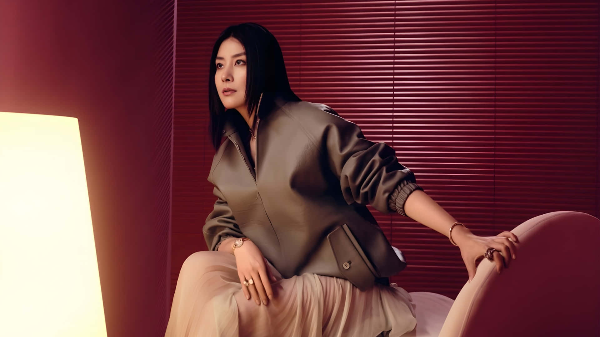  壁紙 — 一位身穿時尚西裝外套的優雅女士，坐在一張弧形椅子上，背景是深紅色的條紋牆，沐浴在溫暖的環境光中。