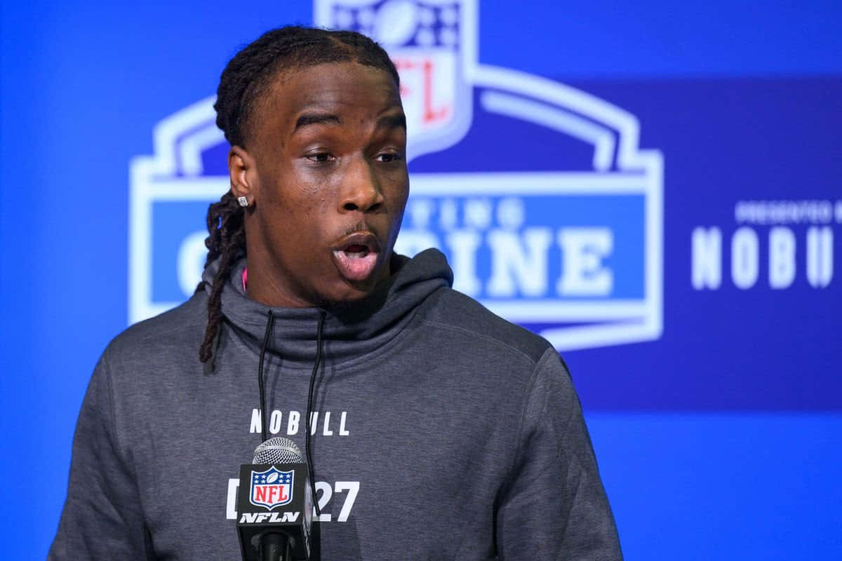  壁紙 — 一位身穿灰色連帽衫的年輕黑人美式足球運動員在NFL聯合試訓記者會上，對著藍色品牌背景的麥克風發言。