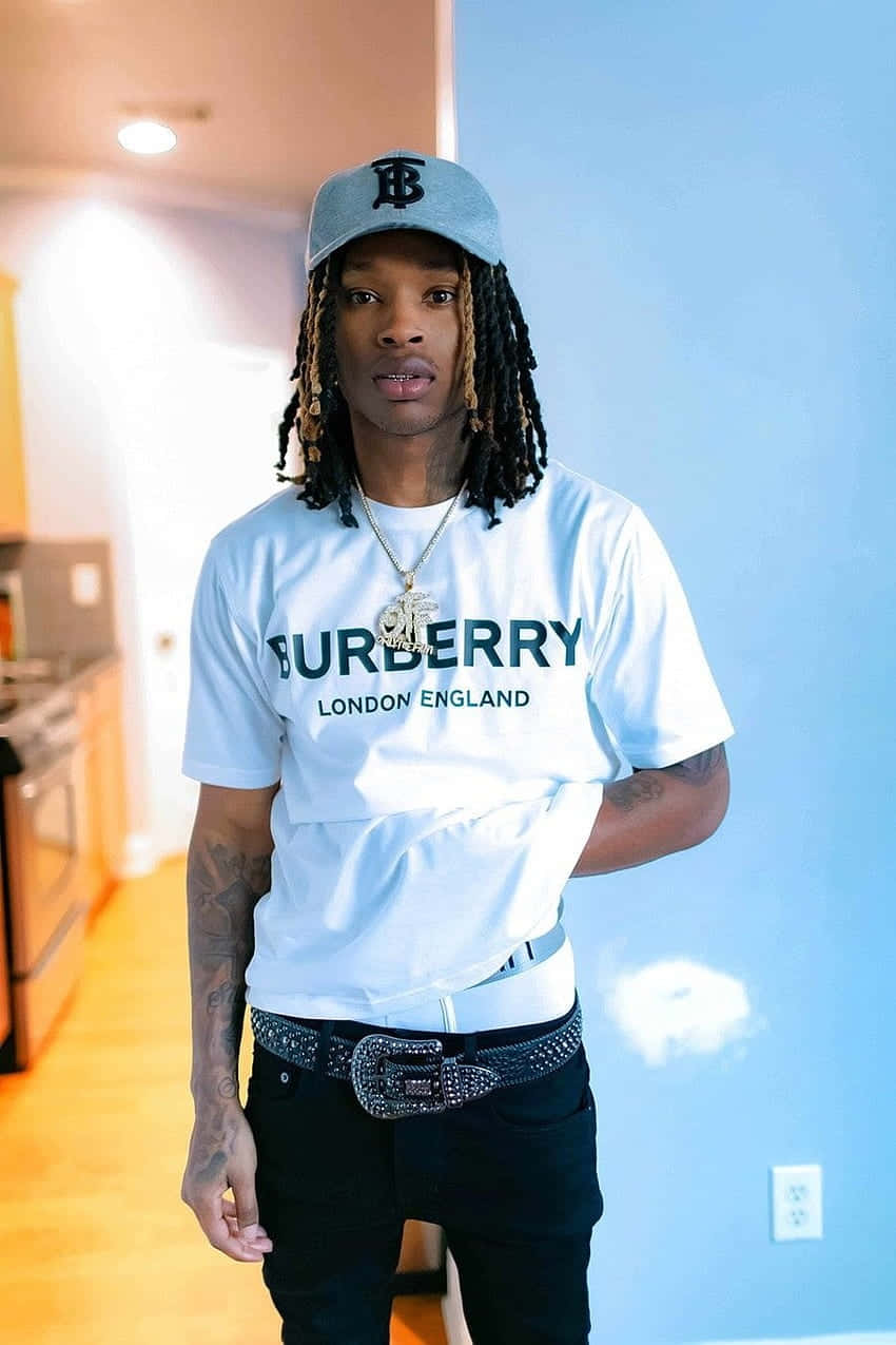  壁紙 — 一位時尚的年輕男子留著辮子，穿著Burberry T恤和鉚釘腰帶，站在藍色牆壁和溫暖燈光的現代廚房裡。