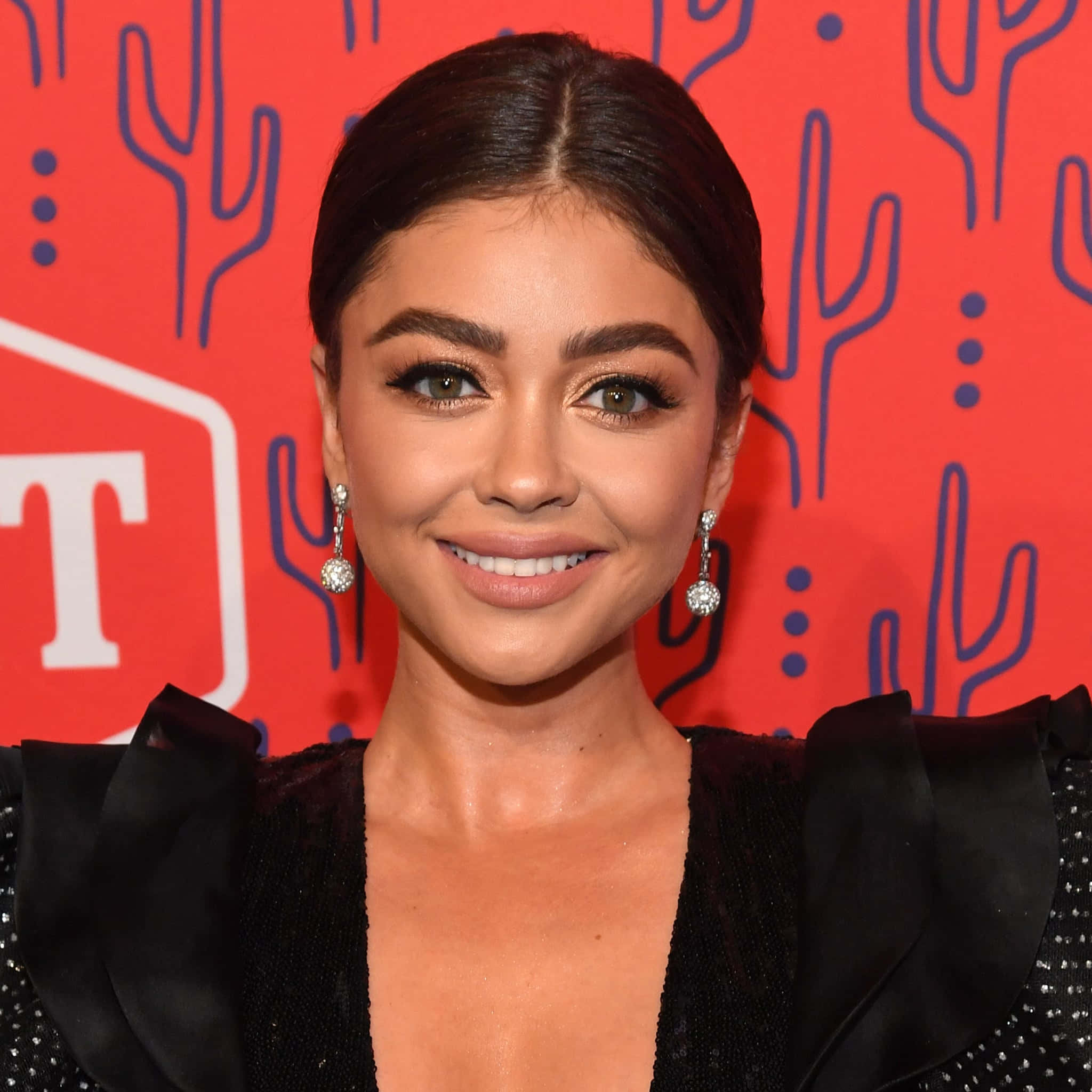 Sarah Hyland 桌布 壁紙 — 一位微笑的黑髮女子戴著閃亮的耳環，在帶有藍色仙人掌圖案的鮮紅色背景前擺姿勢。