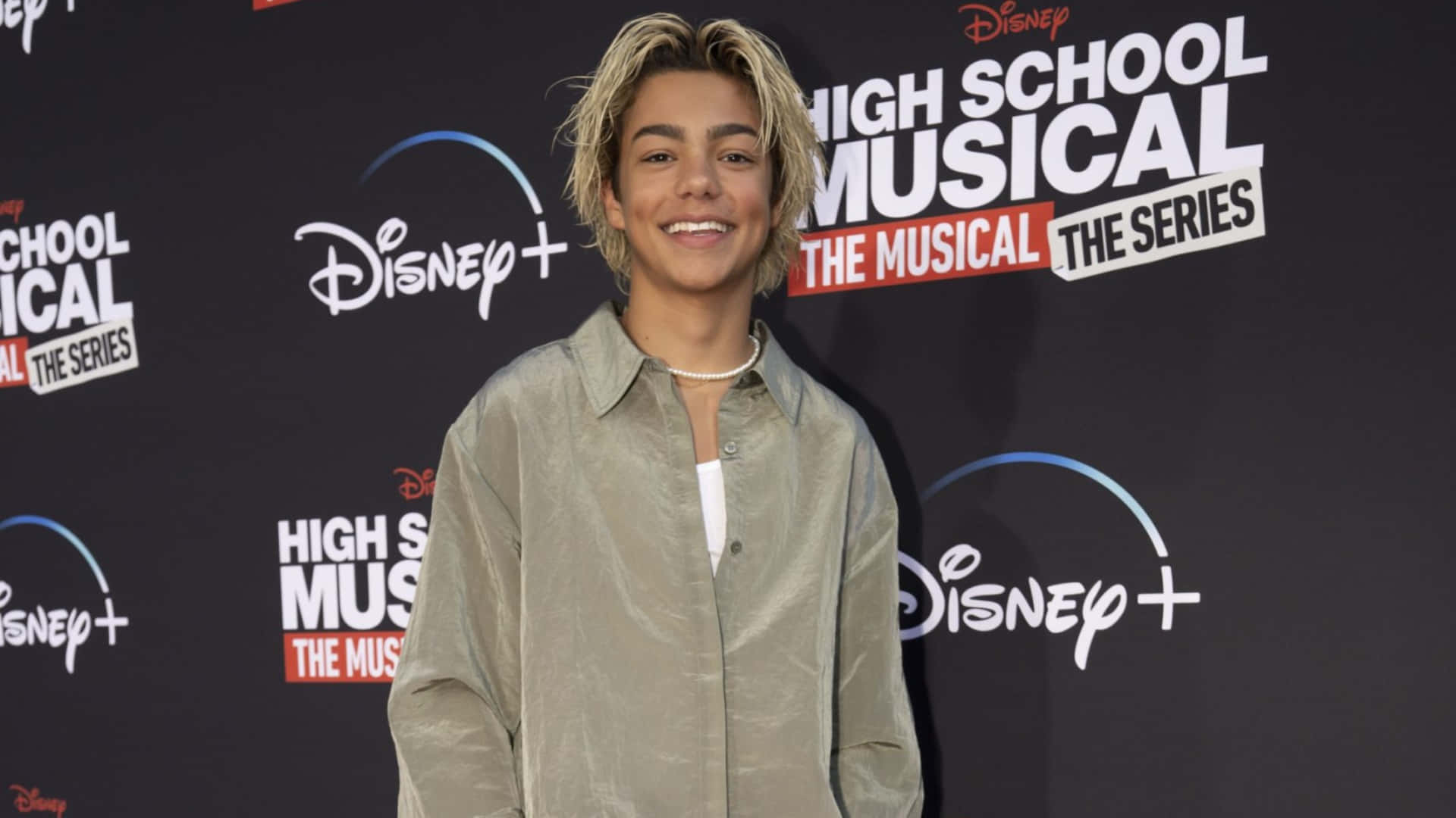  壁紙 — 一位微笑的金髮年輕男子穿著米色襯衫在Disney+ High School Musical The Series活動中擺姿勢。