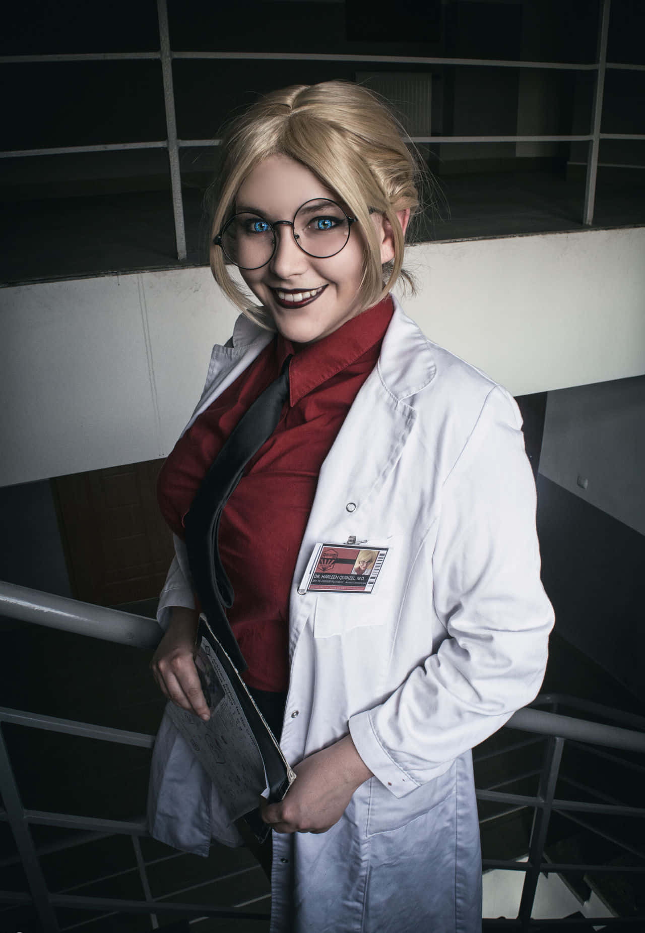 Harleen Quinzel 桌布 壁紙 — 一位微笑的金髮女性，身穿白色實驗袍和紅色襯衫，戴著眼鏡和名牌，自信地在金屬樓梯上擺姿勢。