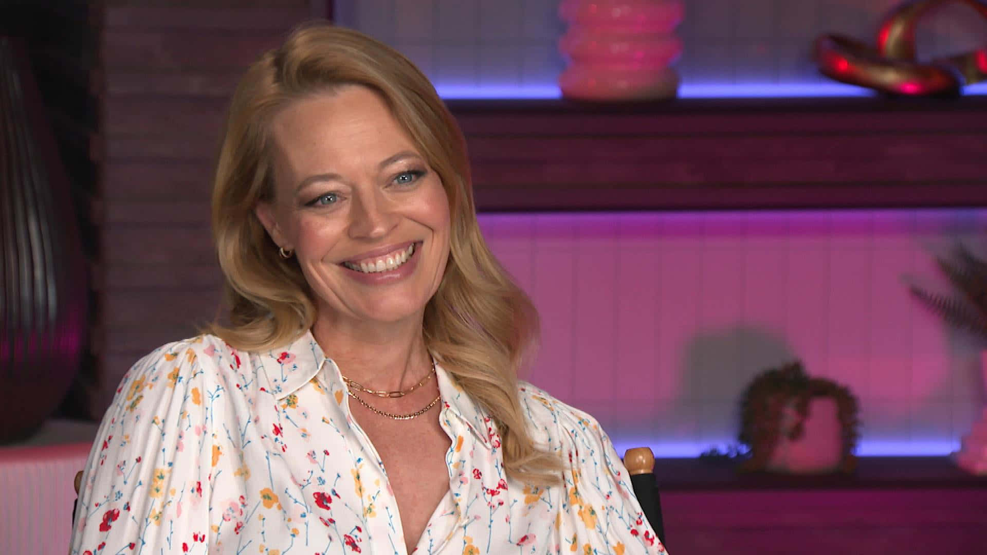 Jeri Ryan 桌布 壁紙 — 一位微笑的金髮女子穿著花卉襯衫，在紫色發光的架子背景下溫暖地坐著。
