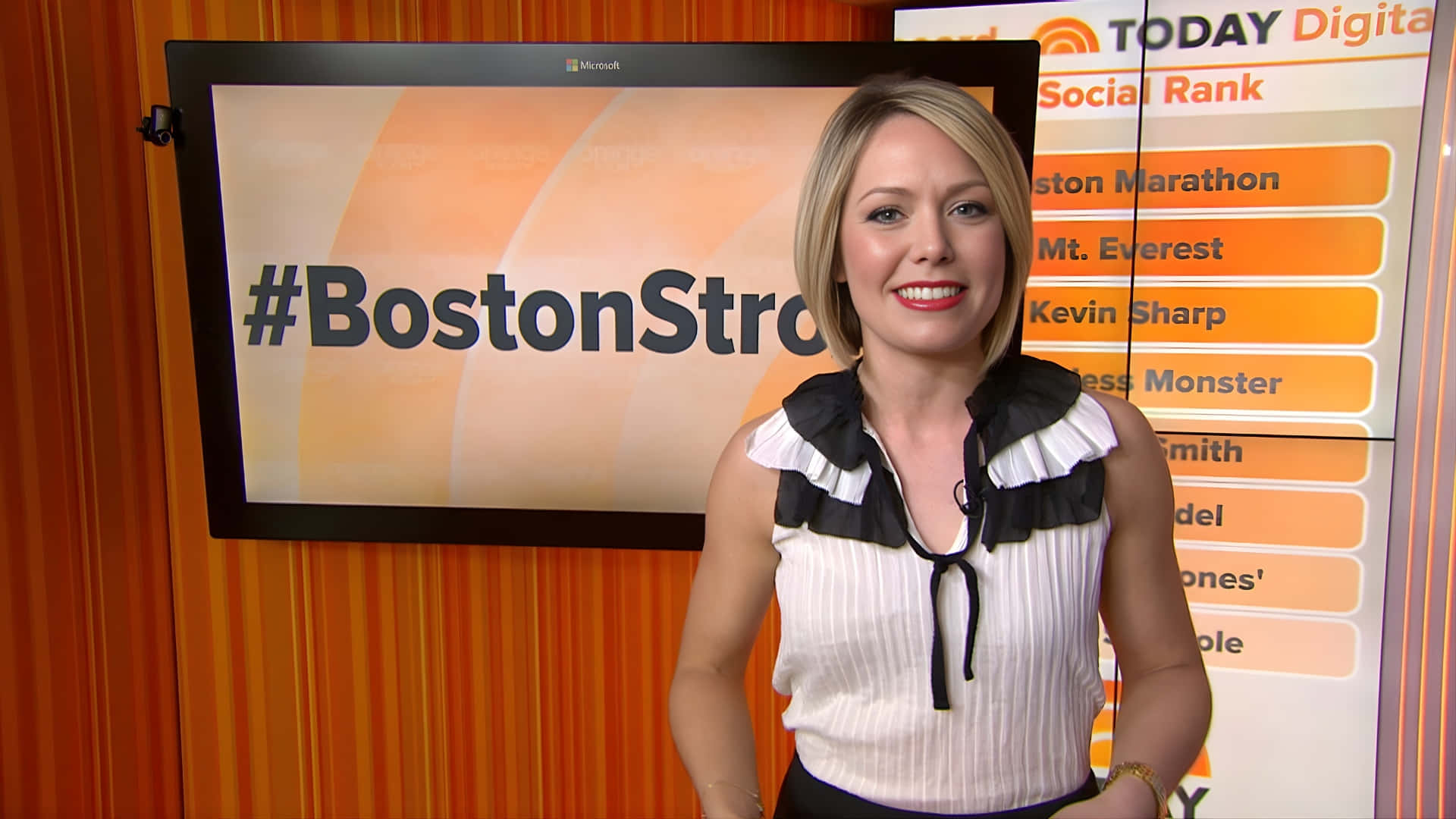 壁紙 — 一位微笑的金髮女主播穿著白色荷葉邊上衣，站在顯示#BostonStrong和社交排名圖形的螢幕前。