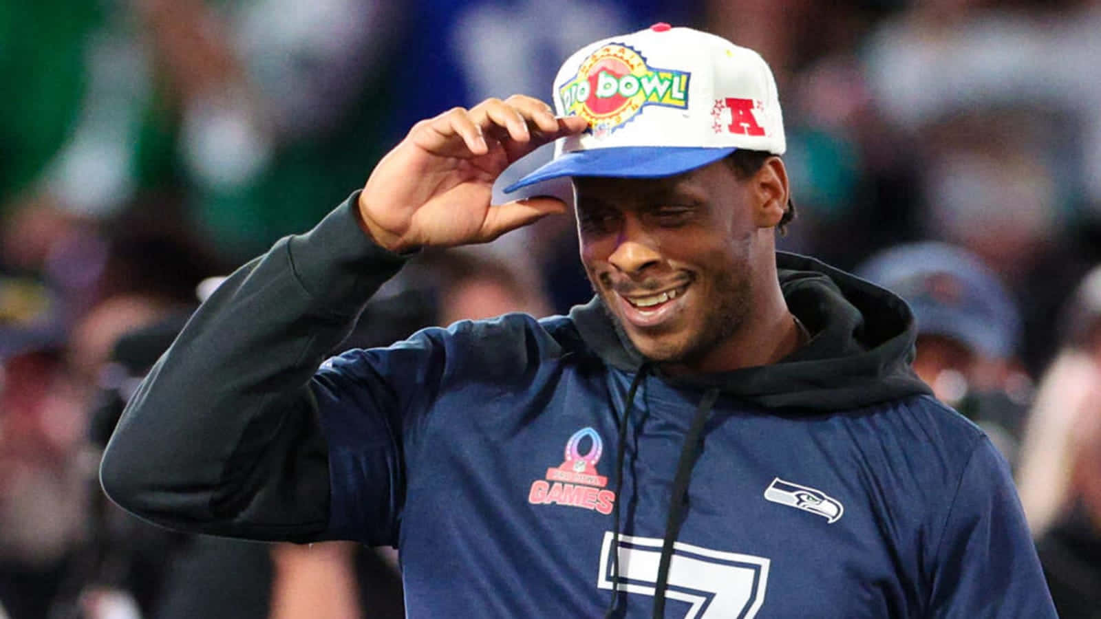  壁紙 — 一位微笑的美式足球員身穿藍色連帽衫，戴著「Pro Bowl」帽子，站在球場上，身後是模糊的體育場人群，正在調整帽子。