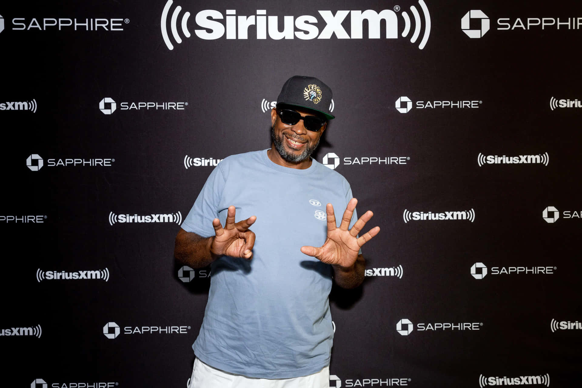  壁紙 — 一位微笑的男子身穿藍色T恤和帽子，雙手張開，在黑色SiriusXM品牌背景前擺姿勢。
