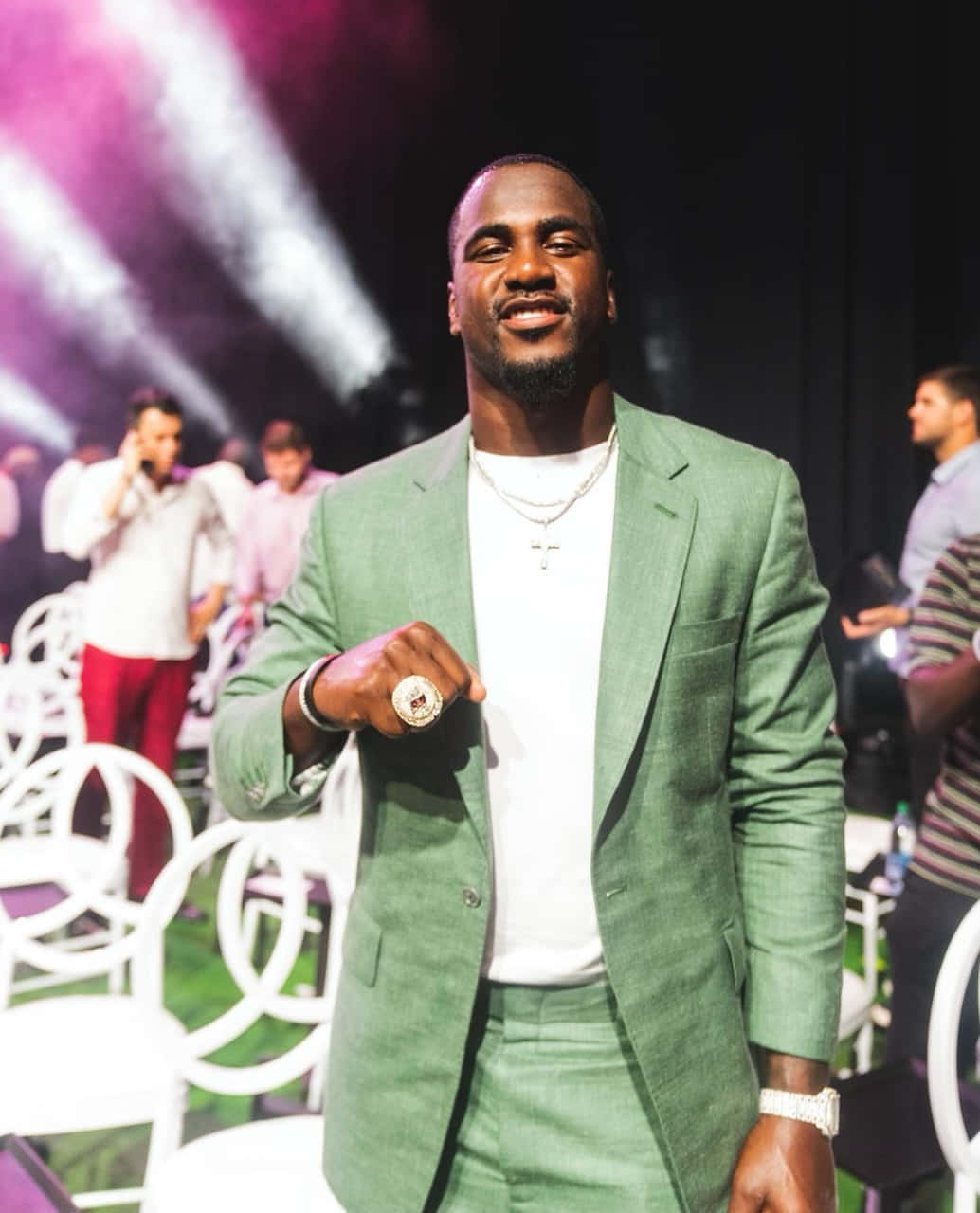  壁紙 — 一位微笑的男子身穿綠色西裝和白色襯衫，在紫色舞台燈光和白色椅子的活力活動中自信地擺姿勢。