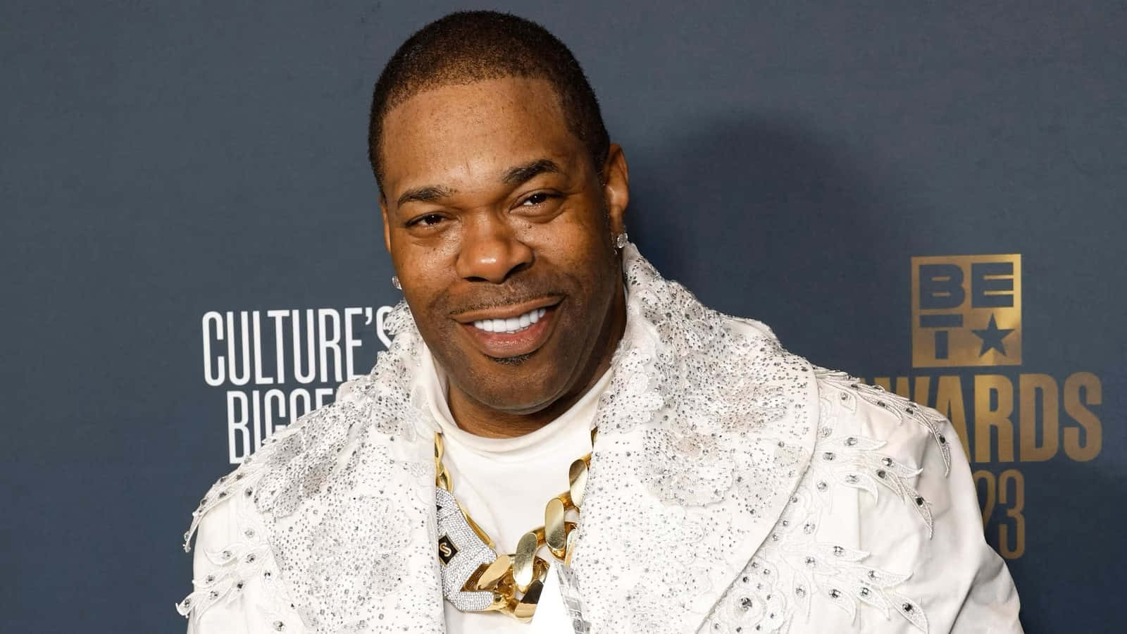  壁紙 — 一位微笑的男子身穿閃亮白色外套和金色鏈條，在BET Awards 2023紅地毯上擺姿勢。
