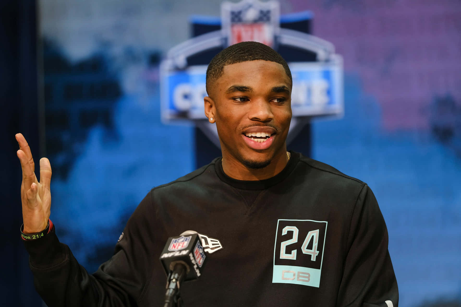  壁紙 — 一位微笑的NFL角衛身穿黑色24號球衣，在記者會上做手勢，身後有麥克風和球隊標誌。