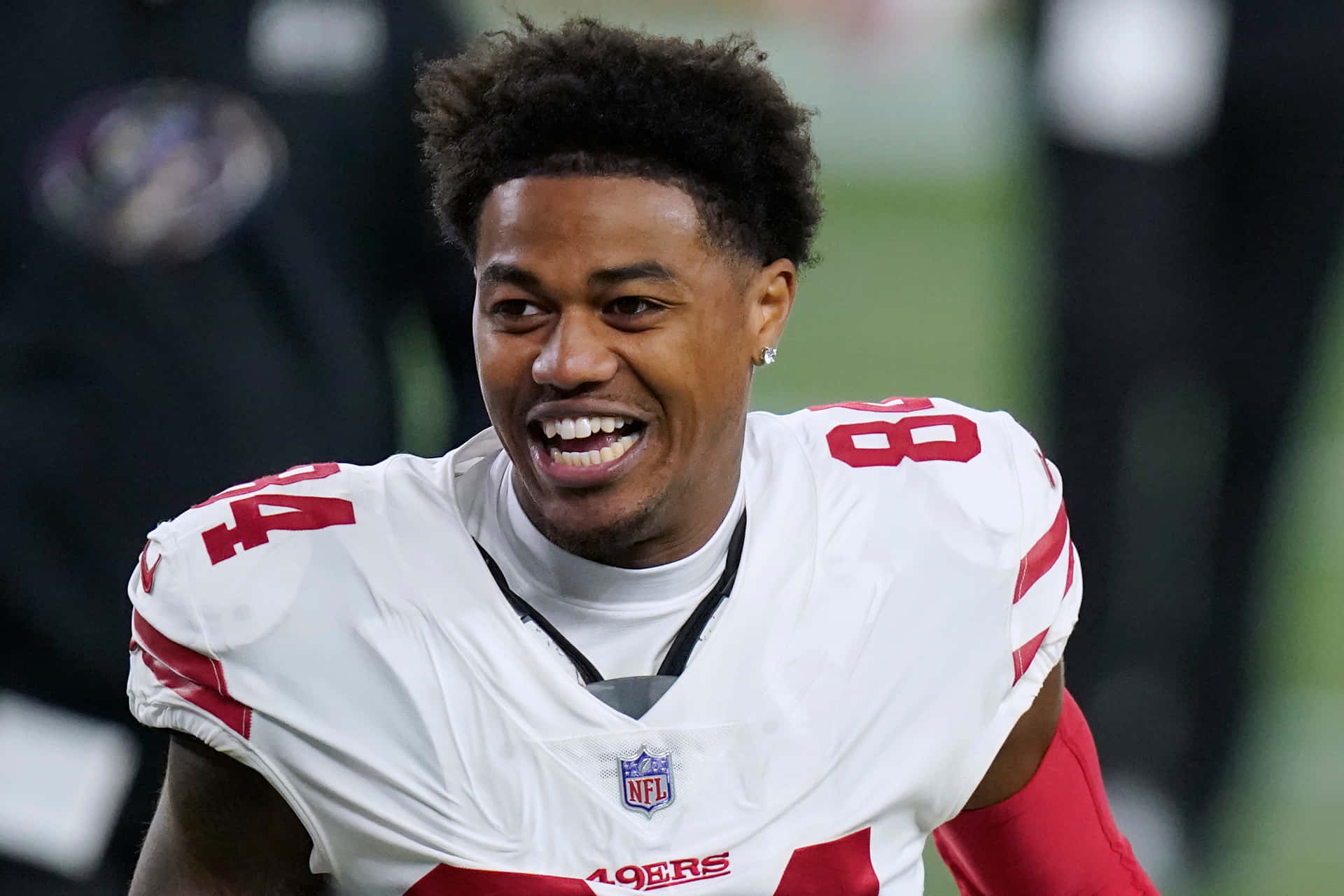  壁紙 — 一位微笑的NFL美式足球員身穿白色和紅色的49ers球衣，在球場上充滿喜悅地慶祝。