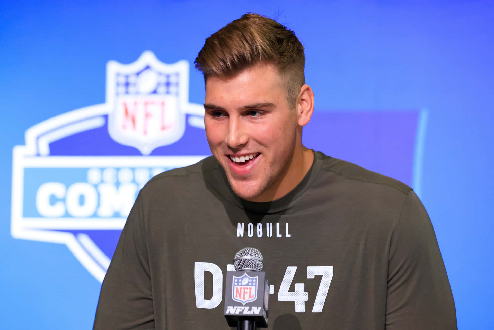  壁紙 — 一位微笑的NFL球員穿著灰色'NOBULL'襯衫，在藍色NFL標誌背景的新聞發布會上對著麥克風講話。