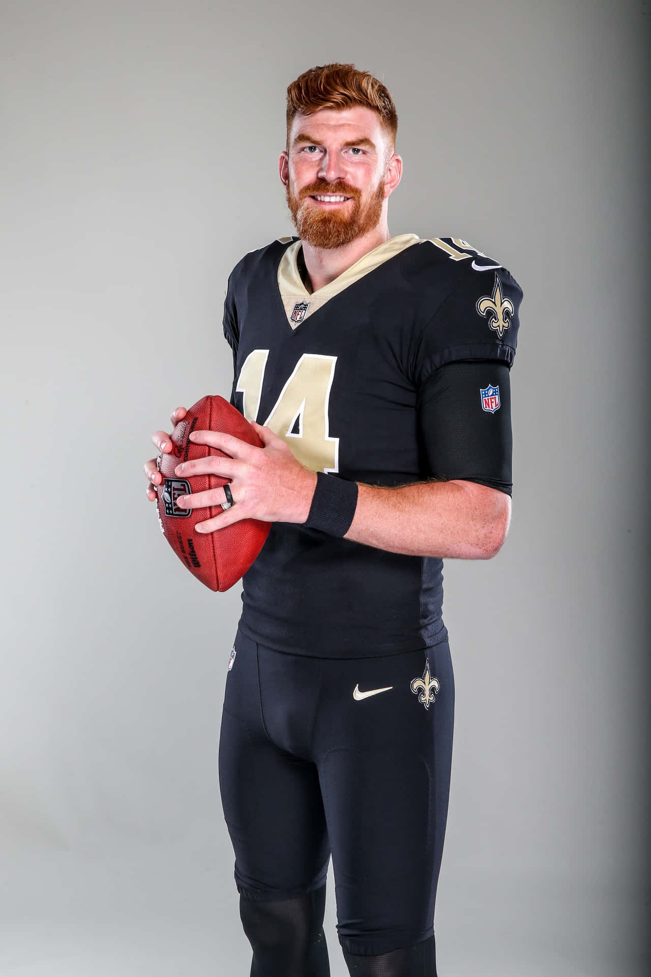  壁紙 — 一位微笑的NFL四分衛穿著黑色和金色的制服，在純灰色工作室背景前拿著一個紅色的美式足球。