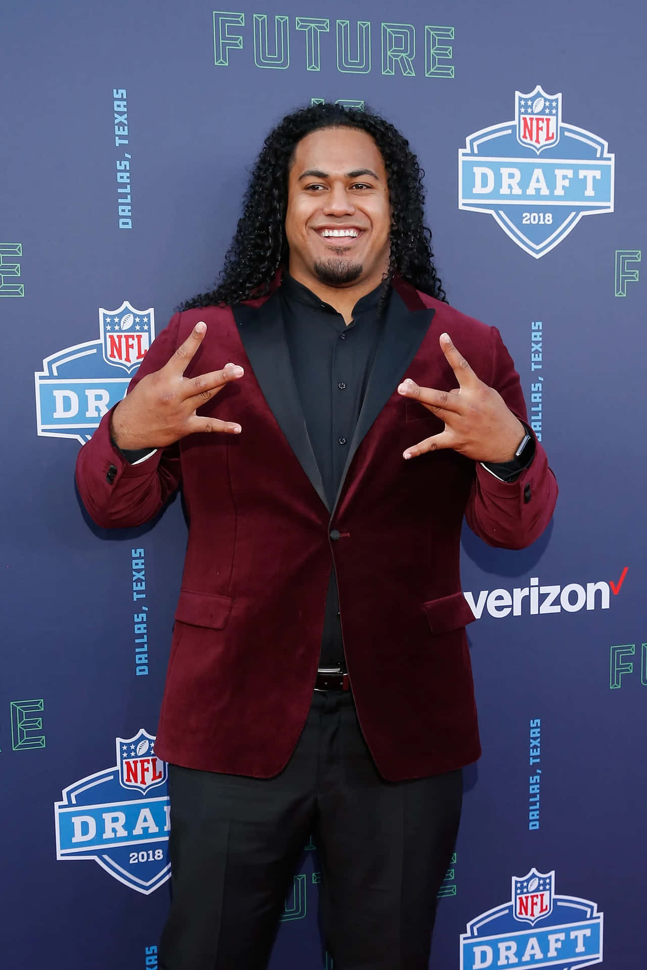  壁紙 — 一位微笑的NFL選秀新秀穿著棗紅色天鵝絨西裝外套，在藍色活動背景前擺出『搖滾手勢』。