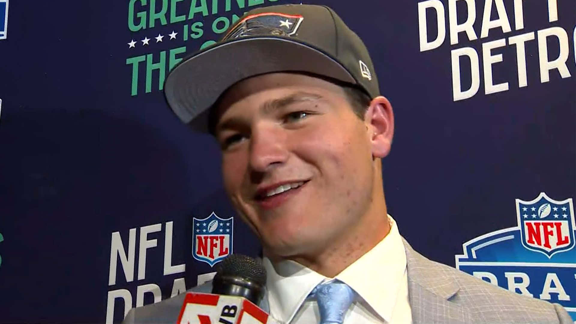  壁紙 — 一位微笑的年輕男子穿著西裝和NFL帽子，在選秀活動中對著麥克風說話。