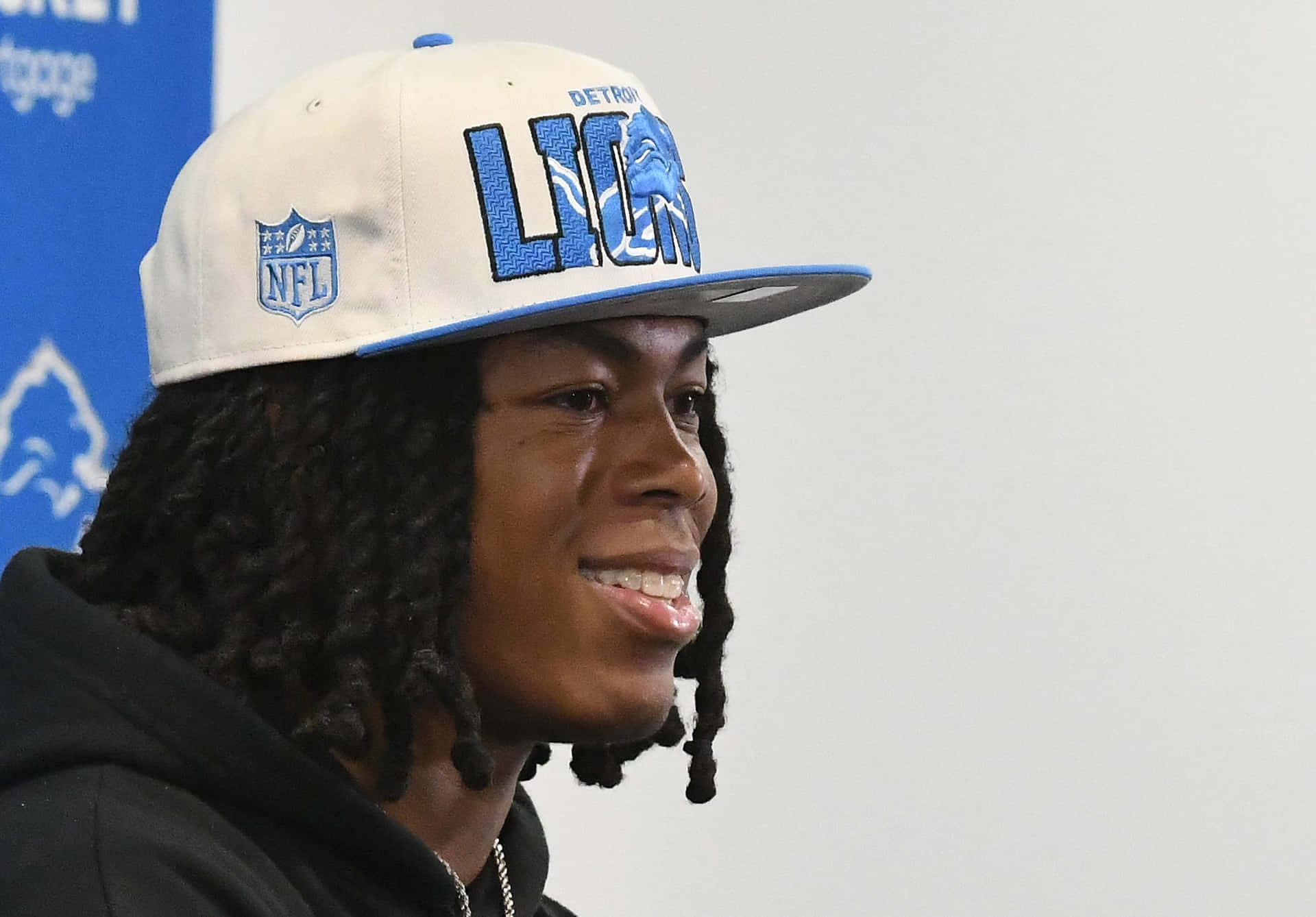  壁紙 — 一位微笑的年輕男子留著辮子，在記者會上戴著底特律雄獅NFL帽子。