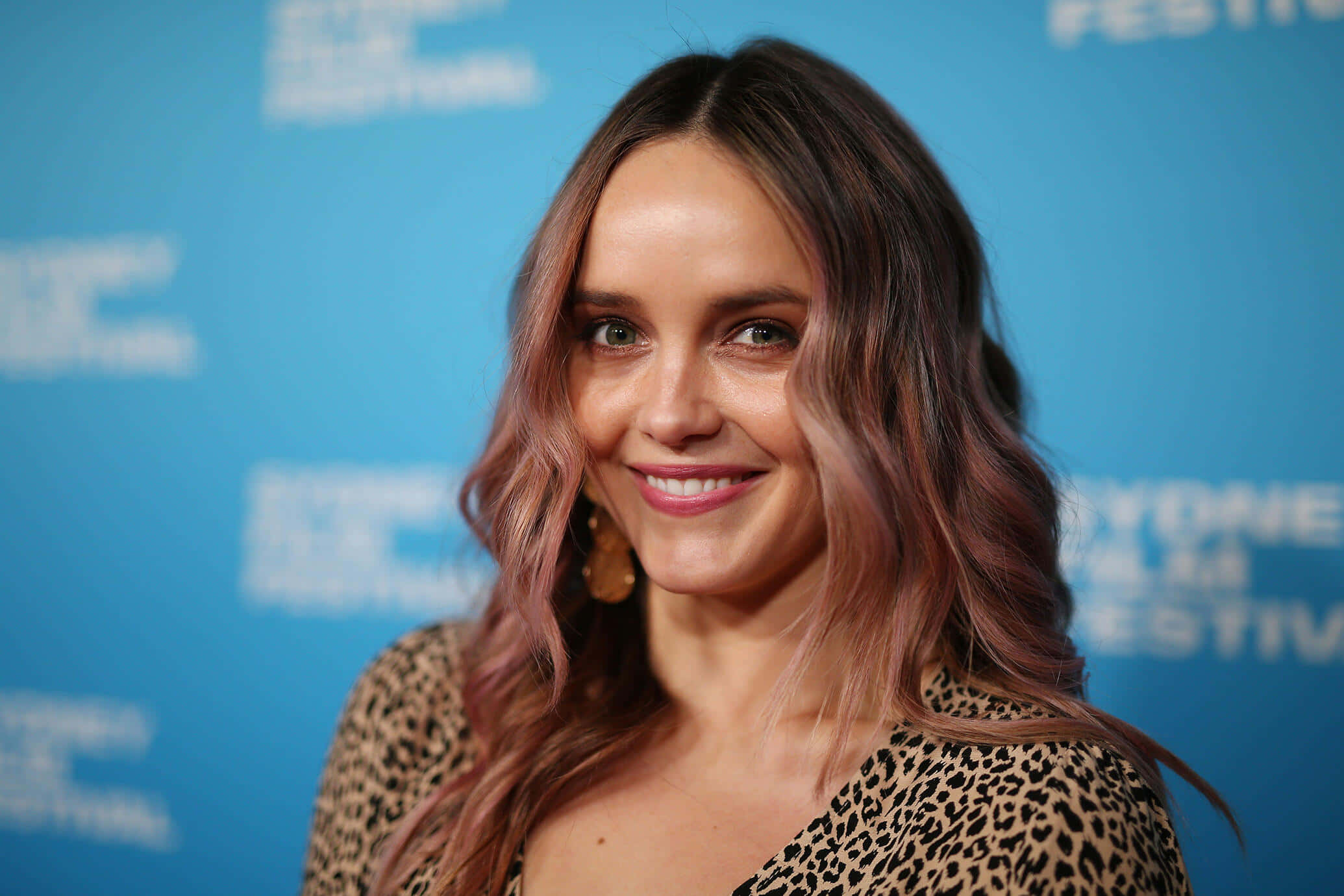 Rebecca Breeds 桌布 壁紙 — 一位微笑的女性，波浪形粉紅色頭髮，穿著豹紋上衣，背景是藍色節日背景。