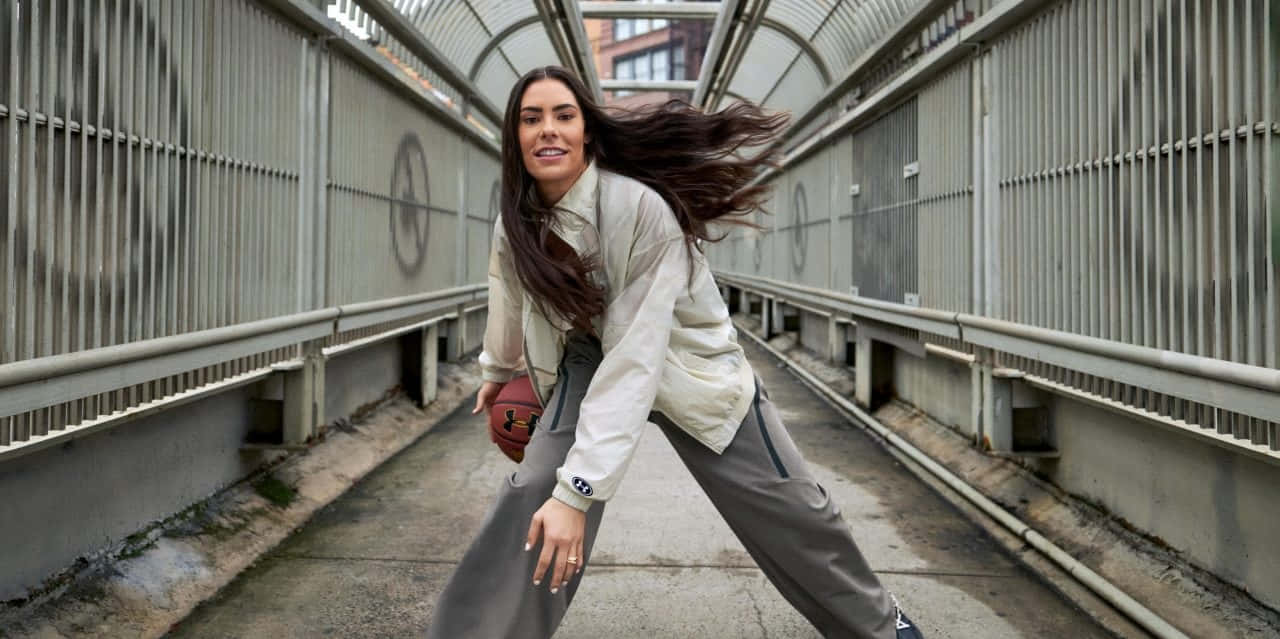  壁紙 — 一位微笑的女性穿著運動服，在金屬步道下運籃球，頭髮飛揚。
