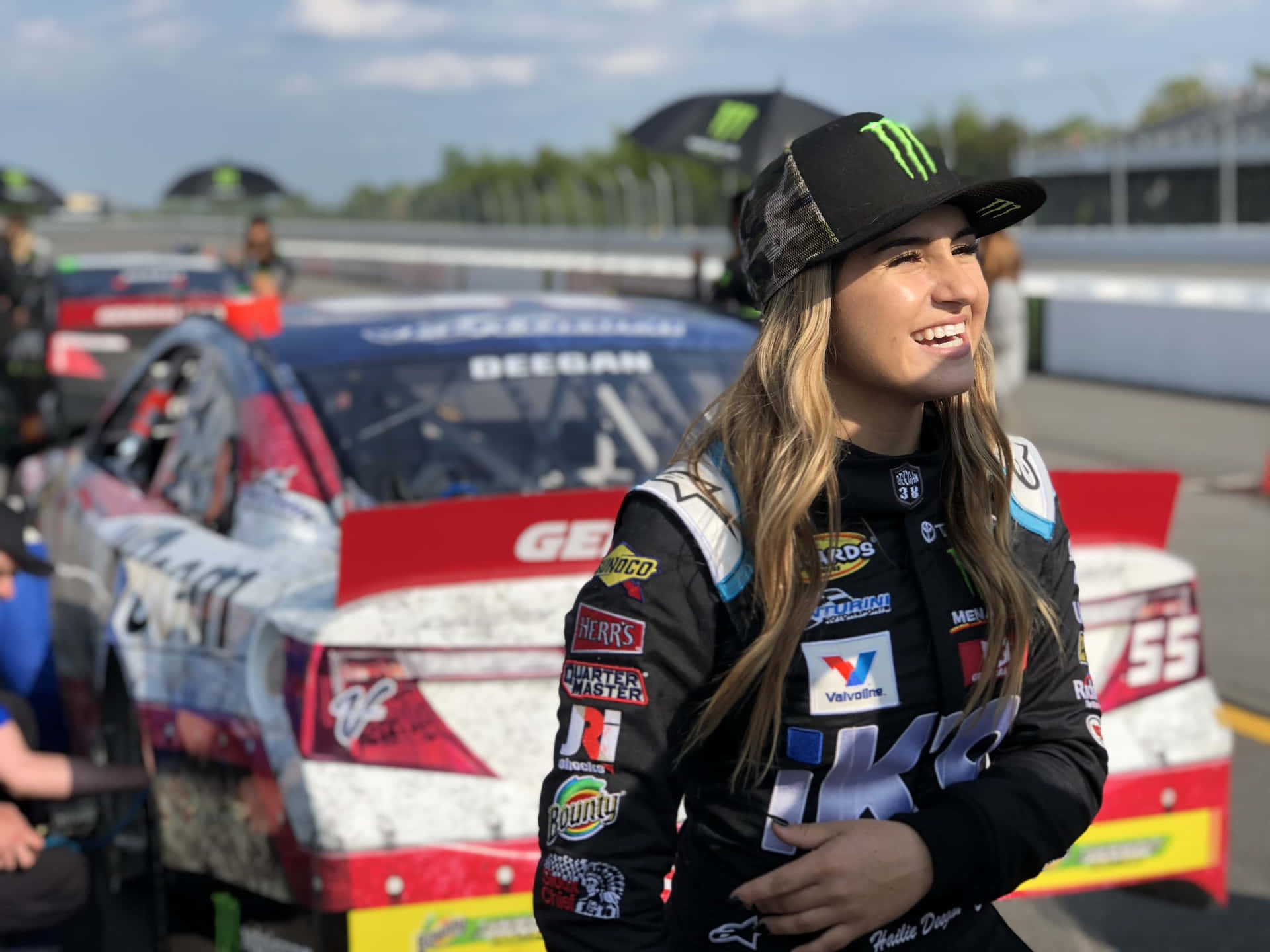  壁紙 — 一位微笑的女性NASCAR車手穿著賽車裝備，站在賽道上她那沾滿泥濘的賽車旁，天空部分多雲。