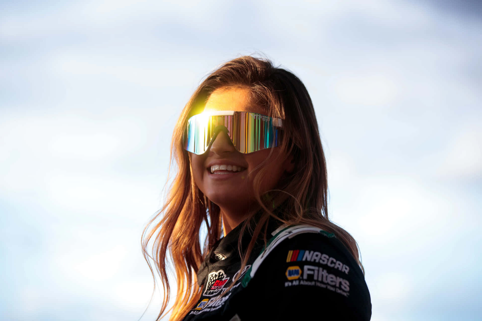  壁紙 — 一位微笑的女性身穿NASCAR賽車服，在明亮的天空下戴著反光太陽眼鏡。