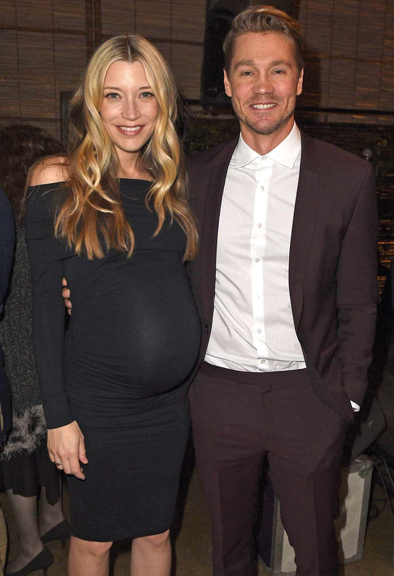 Sarah Roemer 桌布 壁紙 — 一位微笑的孕婦穿著黑色連衣裙，站在穿著栗色西裝的伴侶旁邊，在室內活動中。