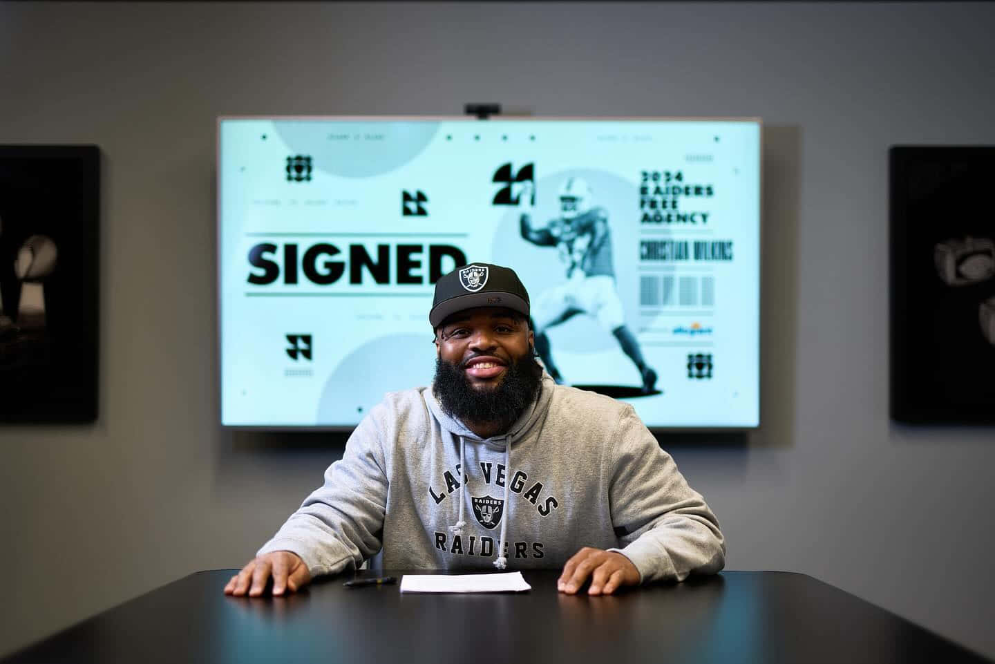  壁紙 — 一位微笑的足球員穿著Raiders連帽衫坐在桌前，身後螢幕顯示「SIGNED」和他的名字。