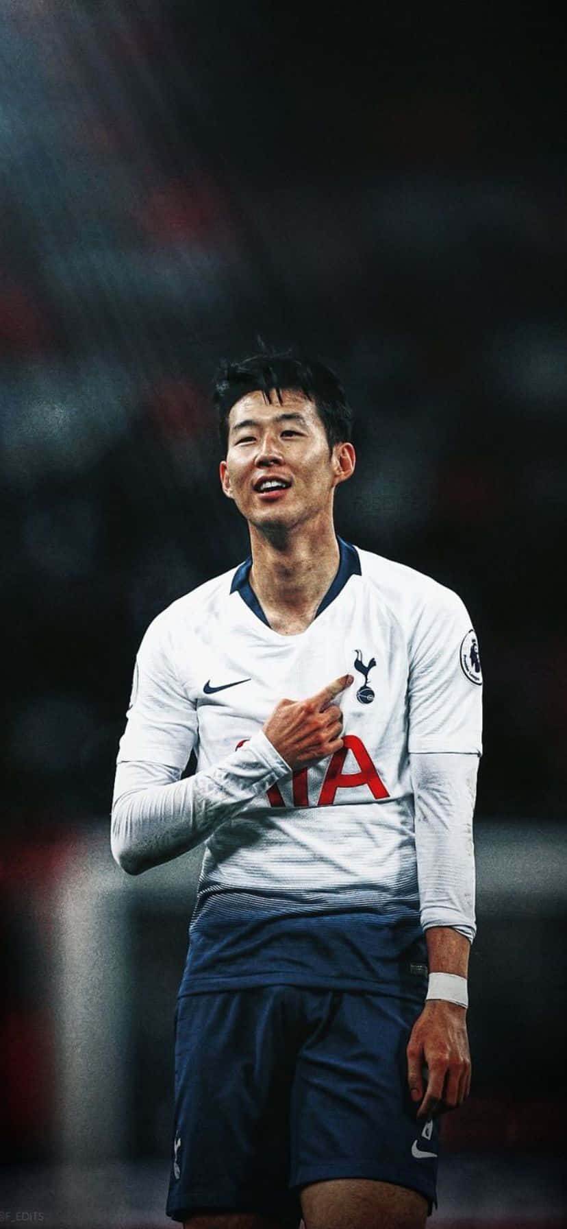  壁紙 — 一位微笑的足球運動員身穿白色熱刺球衣，在球場燈光下自信地指向鏡頭。