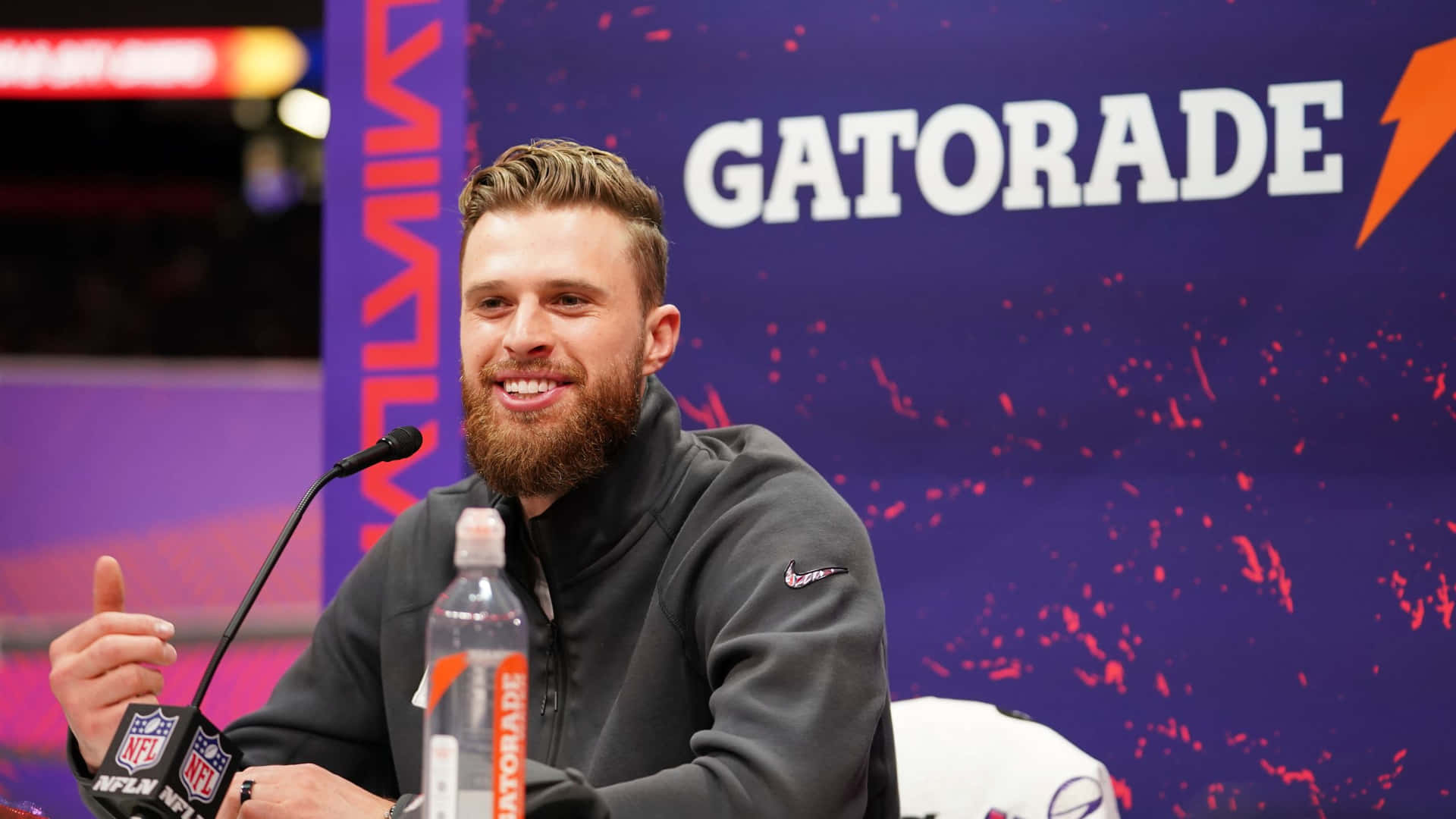  壁紙 — 一位微笑留鬍鬚的男子穿著灰色連帽衫，在帶有NFL標誌的Gatorade品牌記者會上豎起大拇指。