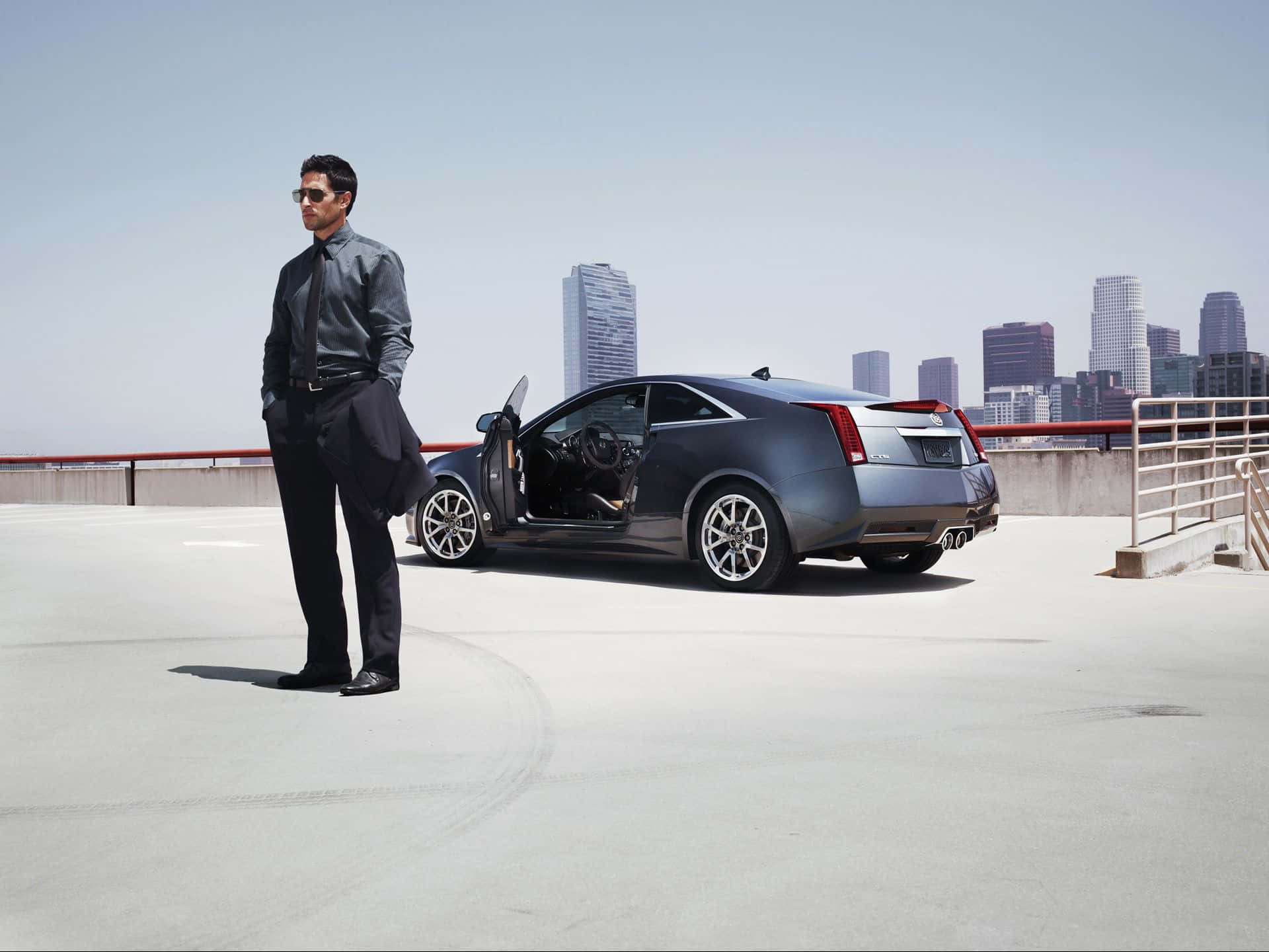 Cadillac Cts 桌布 壁紙 — 一位衣著時尚的男士戴著太陽眼鏡，自信地站在一輛時尚的灰色跑車旁，背景是城市天際線的屋頂上。