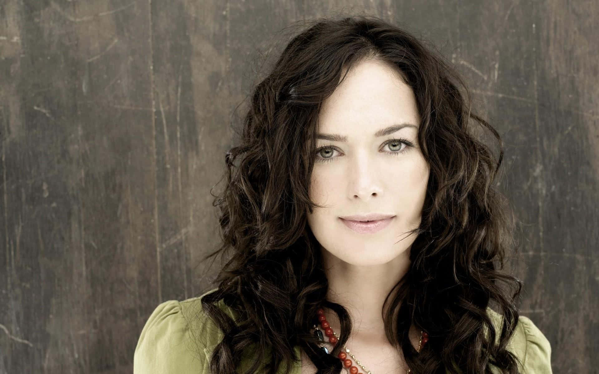 Lena Headey 桌布 壁紙 — 一位擁有深色波浪長髮和綠色眼睛的女性，在風化的木質背景下溫柔地凝視。