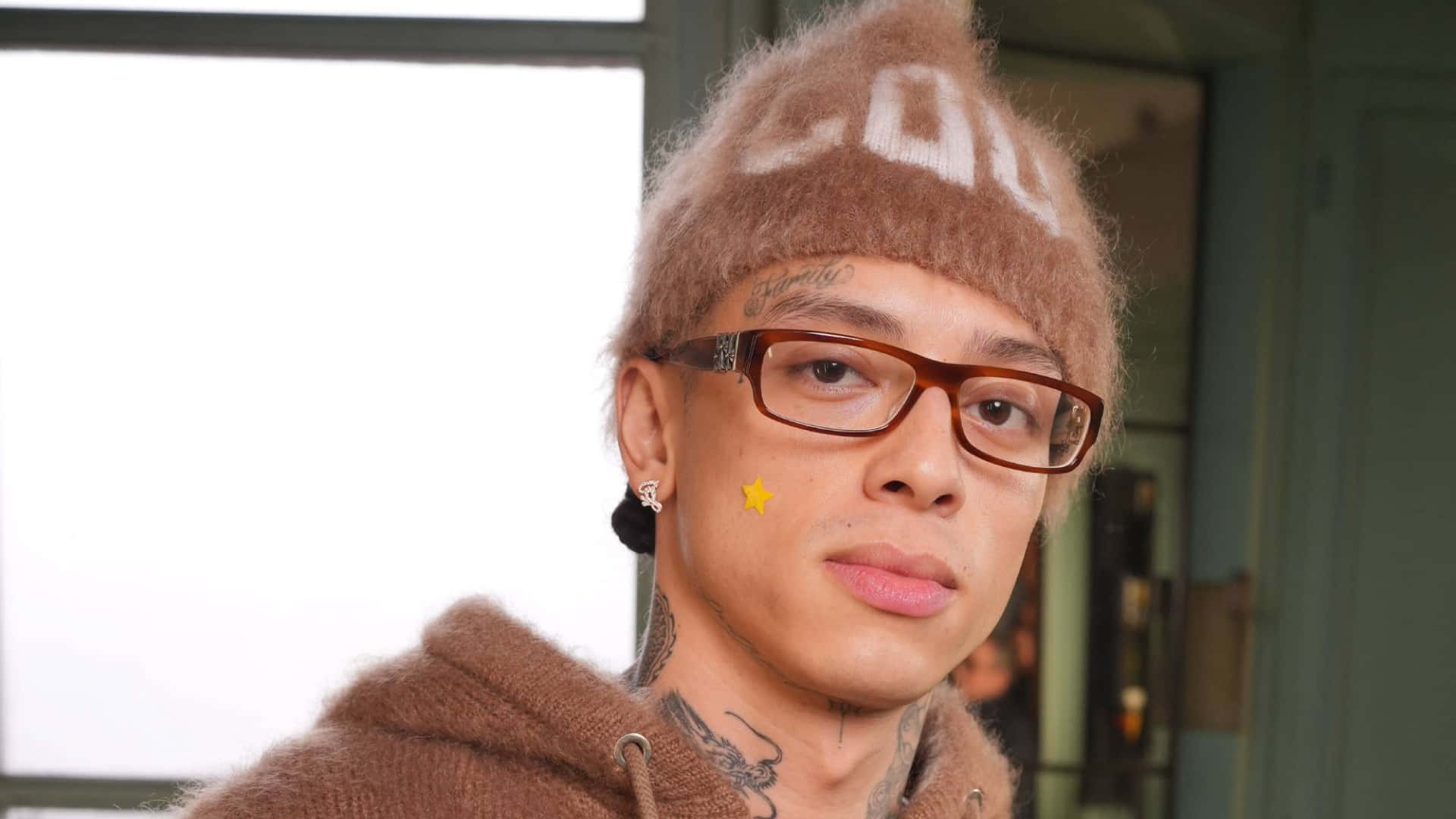  壁紙 — 一位有刺青的年輕男子戴著毛絨棕色毛帽和眼鏡，直視鏡頭。