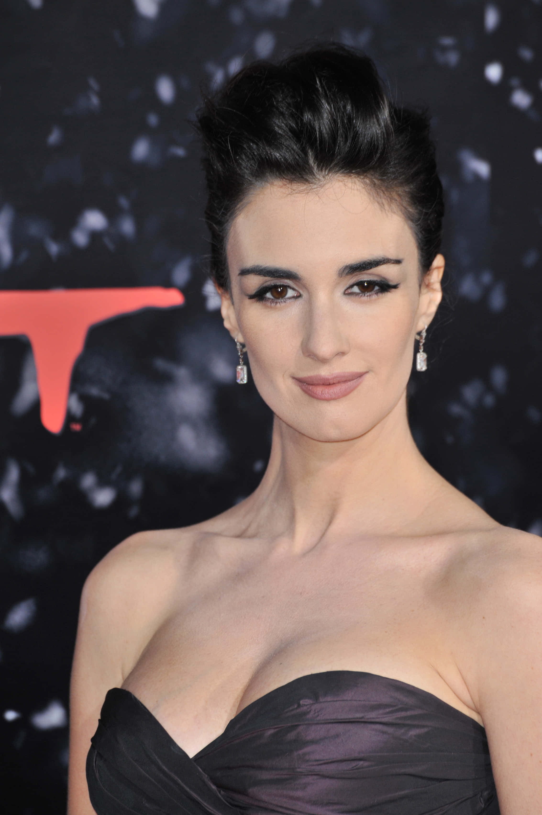 Paz Vega 桌布 壁紙 — 一位優雅的黑髮女子，畫著大膽的眼線，穿著黑色無肩帶禮服，背景是帶有紅色圖案的深色背景。