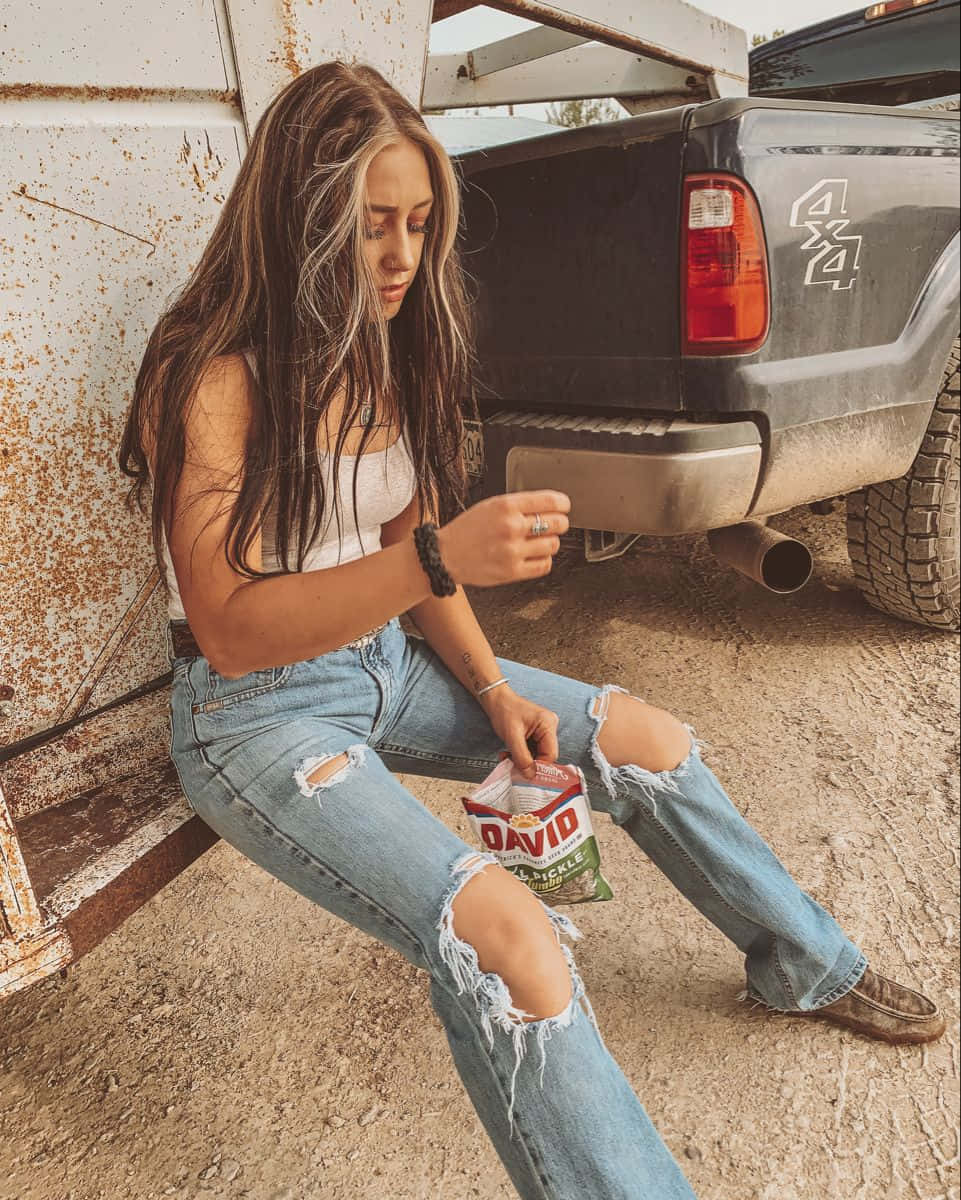  壁紙 — 一位長髮年輕女子坐在生鏽的貨車車斗上，穿著破洞牛仔褲吃薯片。