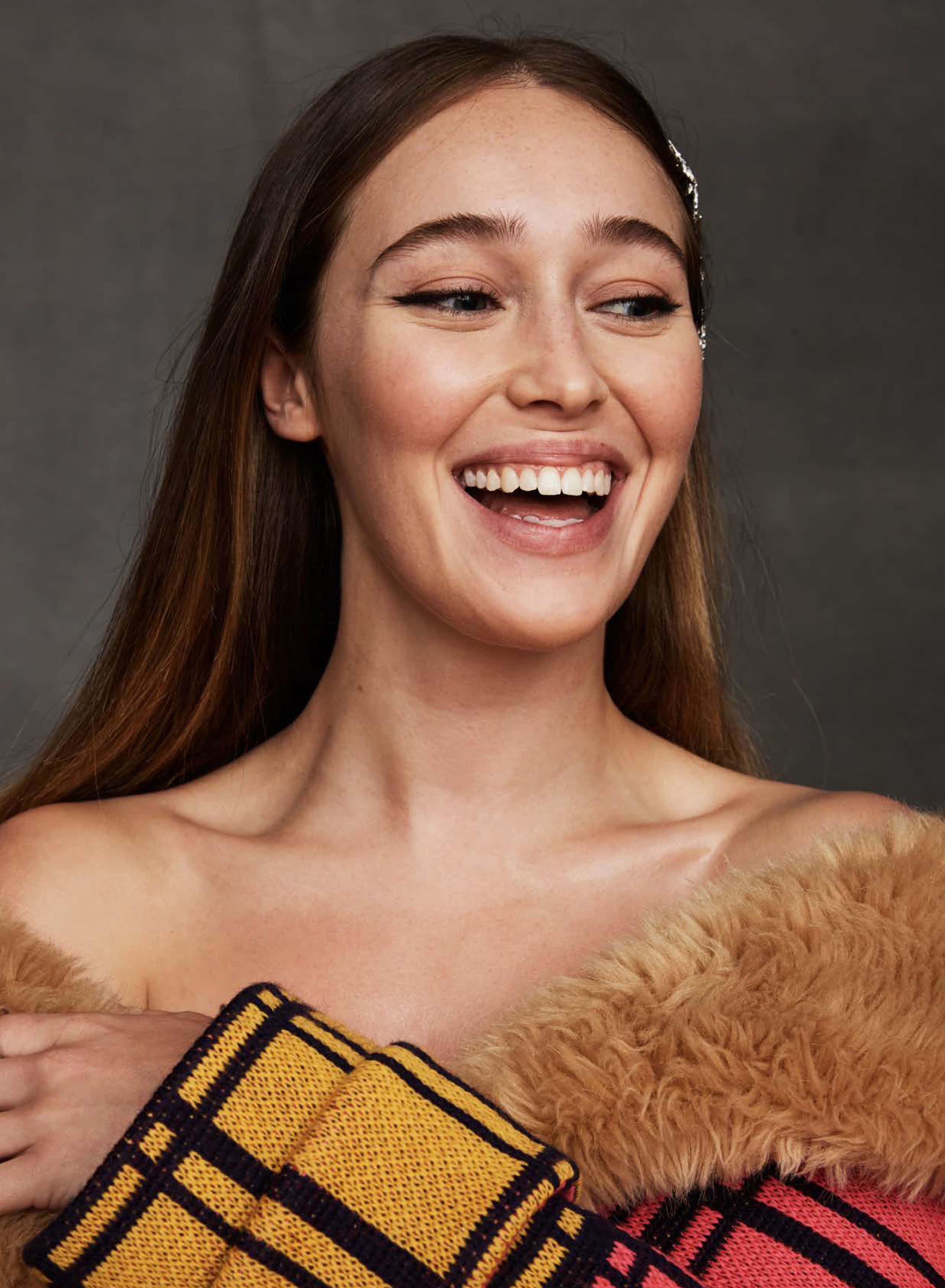 alycia debnam carey 桌布 壁紙 — 一位長著棕色長髮的快樂女性，在灰色背景前，抱著一條帶毛邊的彩色格紋毯，笑得非常燦爛。