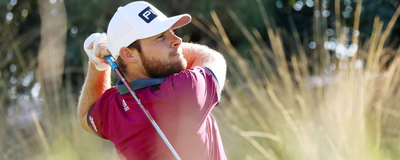 Tyrrell Hatton 桌布 壁紙 — 一位專注的高爾夫球手身穿洋紅色襯衫和白色帽子，在陽光普照的高草背景下執行強力揮桿。