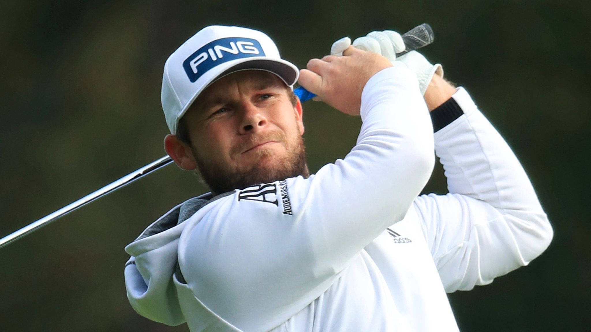 Tyrrell Hatton 桌布 壁紙 — 一位專注的高爾夫球手正在揮桿，戴著白色PING帽子和長袖襯衫，背景是模糊的綠色。