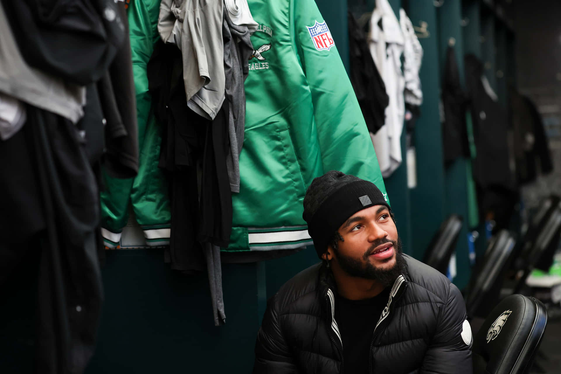  壁紙 — 一位專注的NFL球員戴著黑色毛帽和羽絨外套，坐在被掛著的Eagles裝備包圍的更衣室裡。