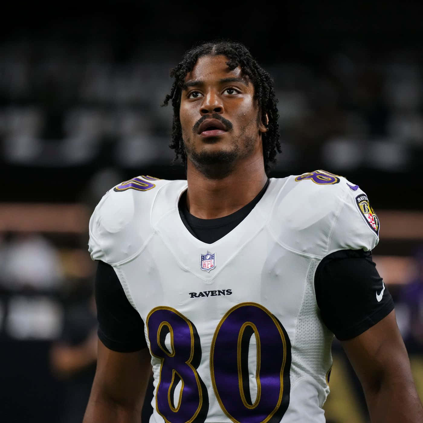  壁紙 — 一位專注的NFL球員，身穿白色和紫色的Ravens 80號球衣，站在球場的燈光下。