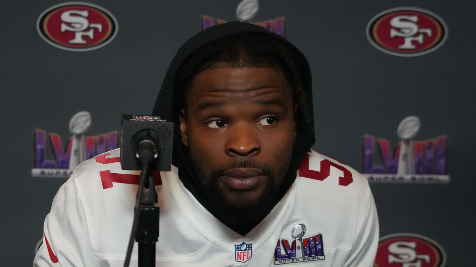  壁紙 — 一位專注的NFL球員身穿白色連帽衫和球衣，坐在記者會麥克風前，身後有超級碗LVIII標誌。