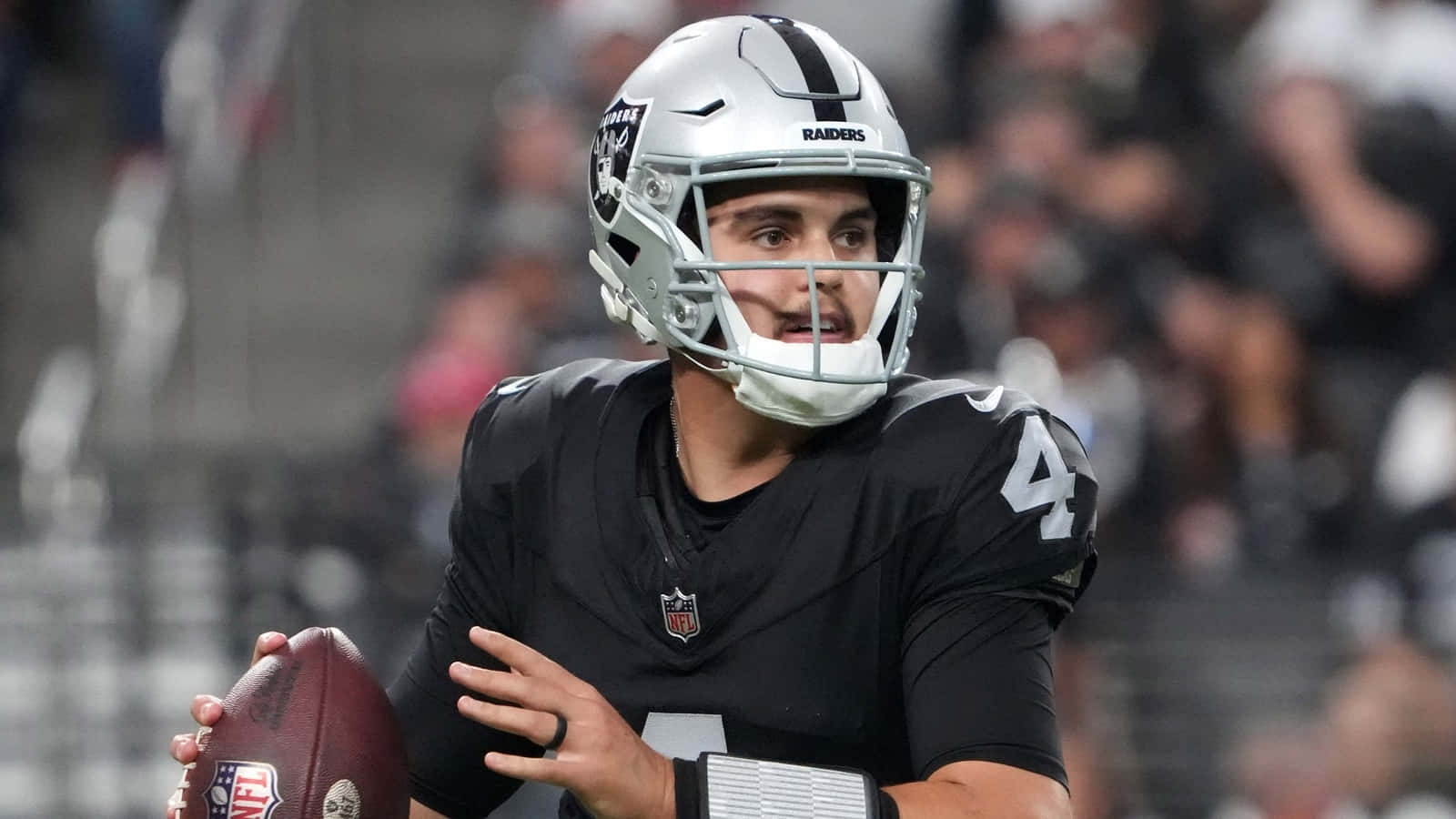  壁紙 — 一位專注的NFL四分衛穿著黑色Raiders球衣，在體育場燈光下準備在比賽中擲出橄欖球。