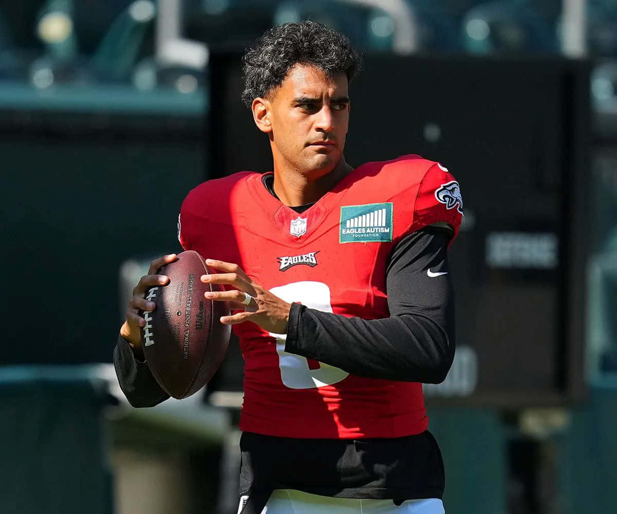  壁紙 — 一位專注的NFL四分衛穿著紅色老鷹隊球衣，在陽光明媚的體育場上持球準備投擲。