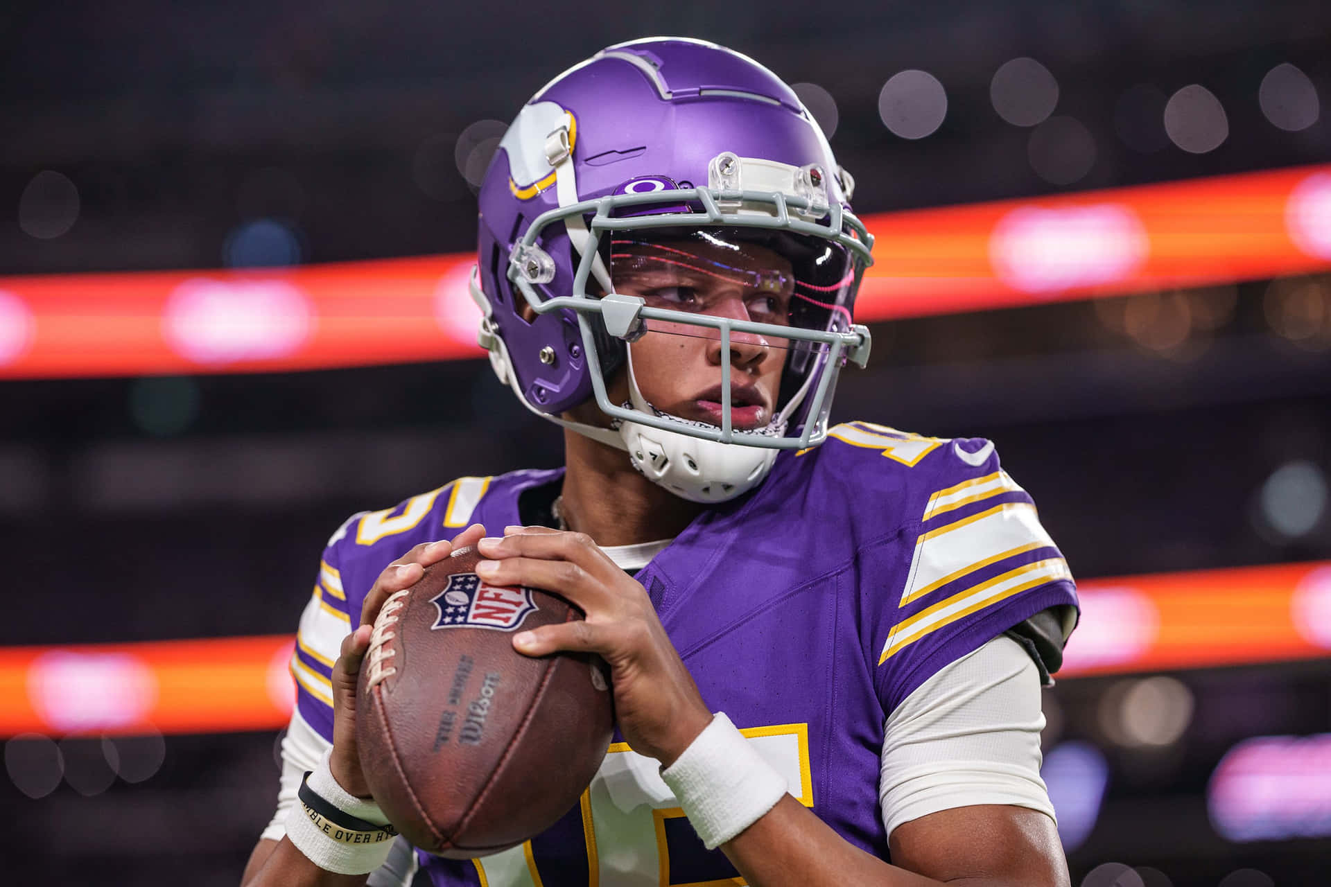  壁紙 — 一位專注的NFL四分衛穿著紫色和金色的裝備，在夜間比賽的體育場燈光下緊握著橄欖球。