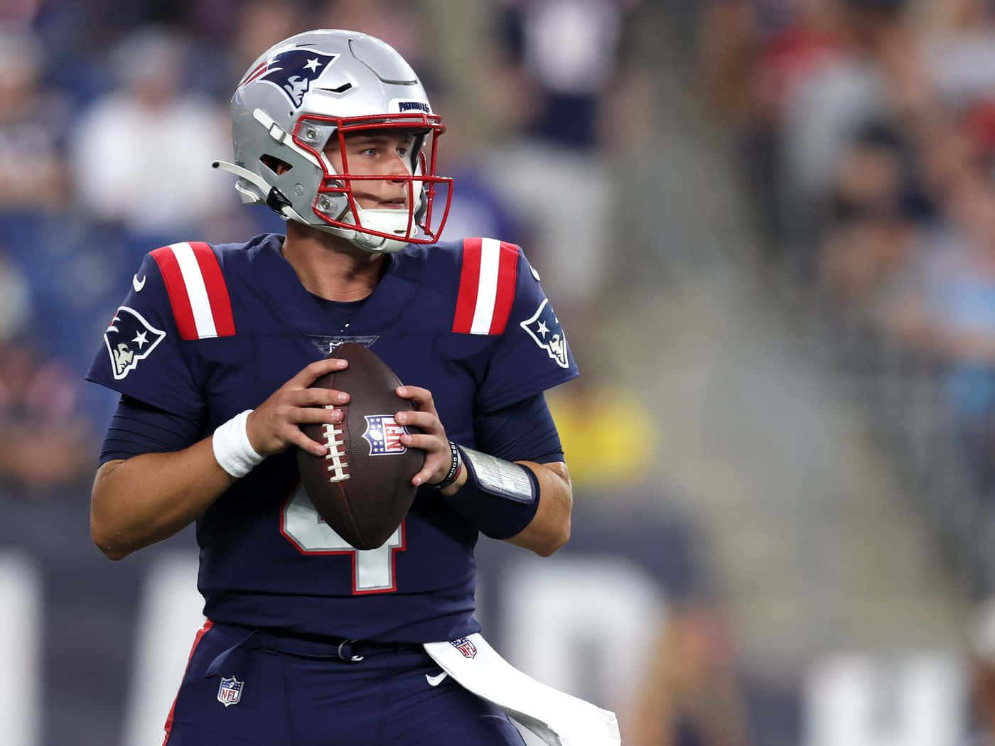  壁紙 — 一位專注的NFL四分衛穿著新英格蘭愛國者隊制服，在體育館燈光下的比賽中持球準備傳球。