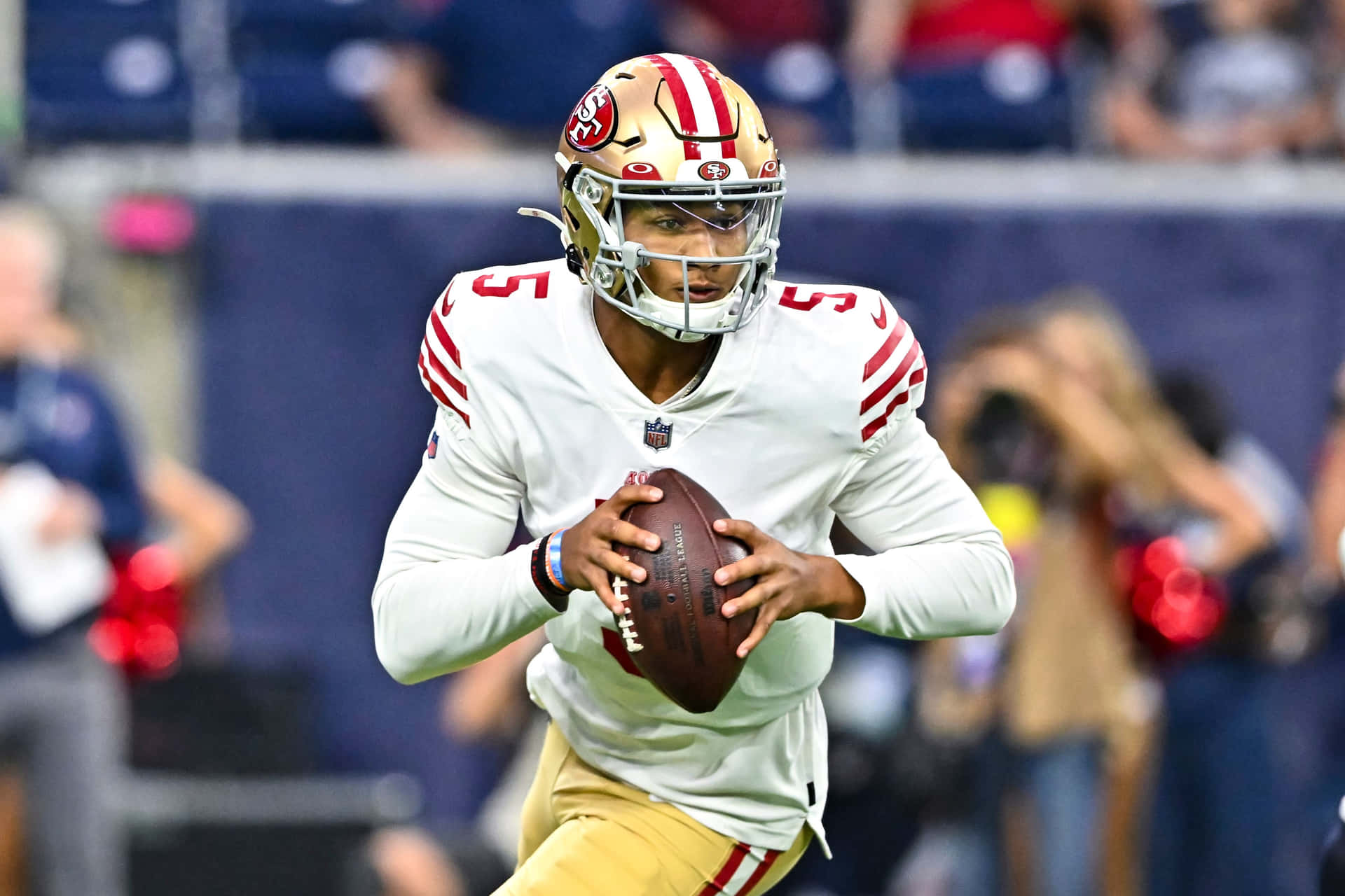  壁紙 — 一位專注的NFL四分衛身穿白色和金色制服，在球場燈光下於比賽中持球。