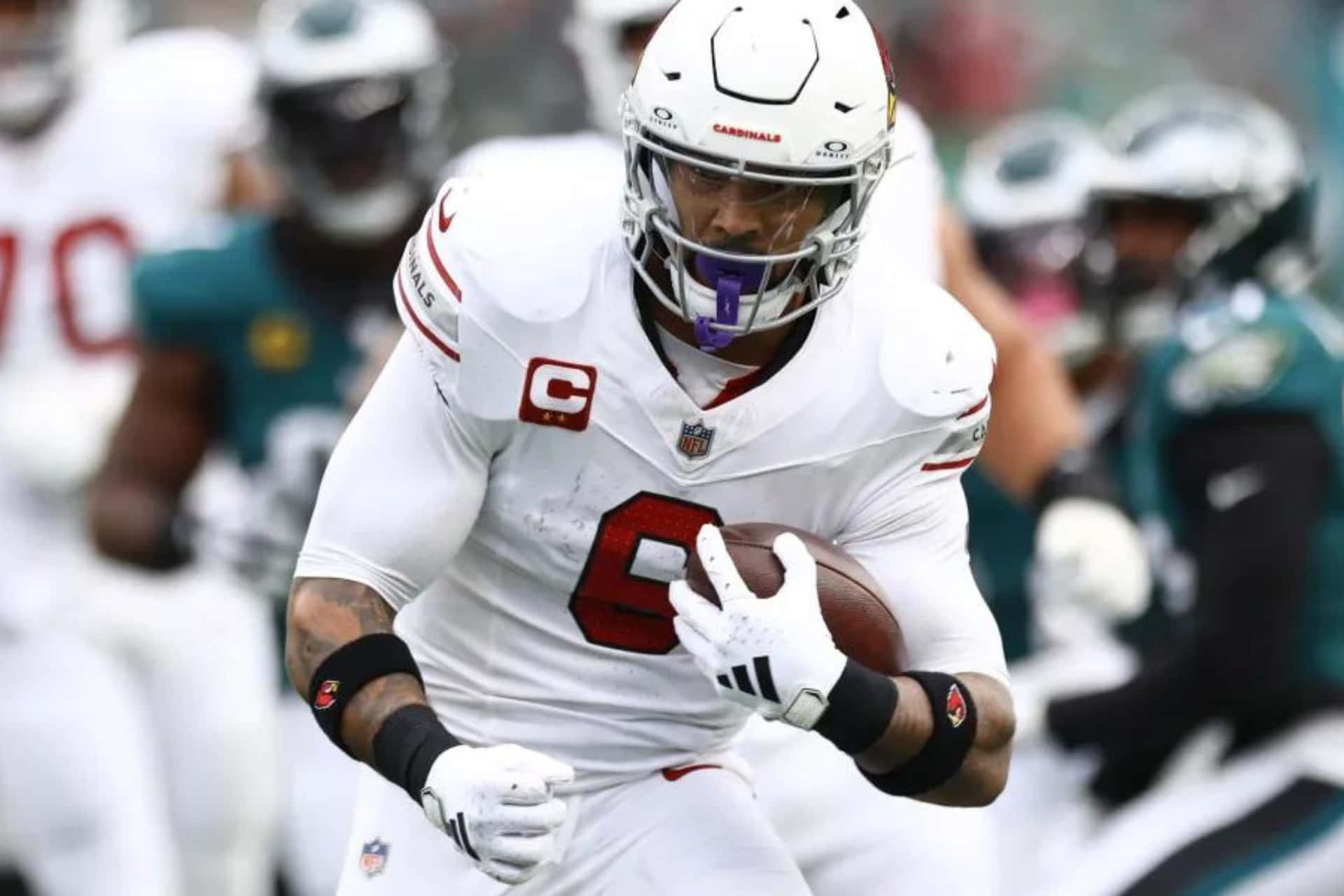  壁紙 — 一位專注的NFL四分衛身穿白色紅雀隊裝備，在日間比賽中帶著橄欖球奔跑，背景對手模糊。