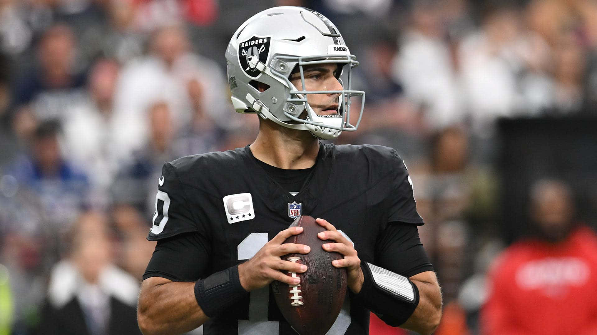  壁紙 — 一位專注的NFL四分衛身穿黑色Raiders球衣，在體育場燈光下抱著橄欖球，背景是模糊的球迷。
