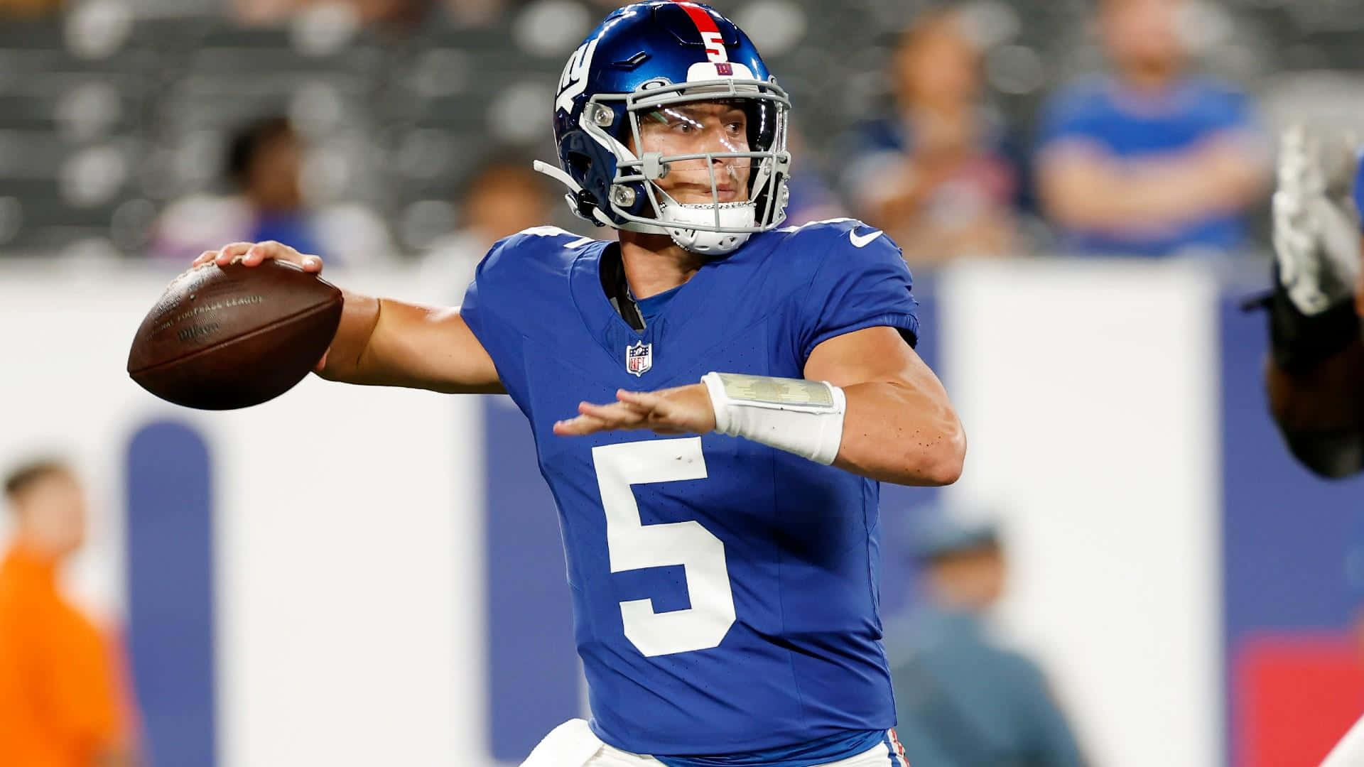  壁紙 — 一位專注的NFL四分衛身穿藍色球衣，號碼5，在體育場燈光下準備擲出橄欖球。
