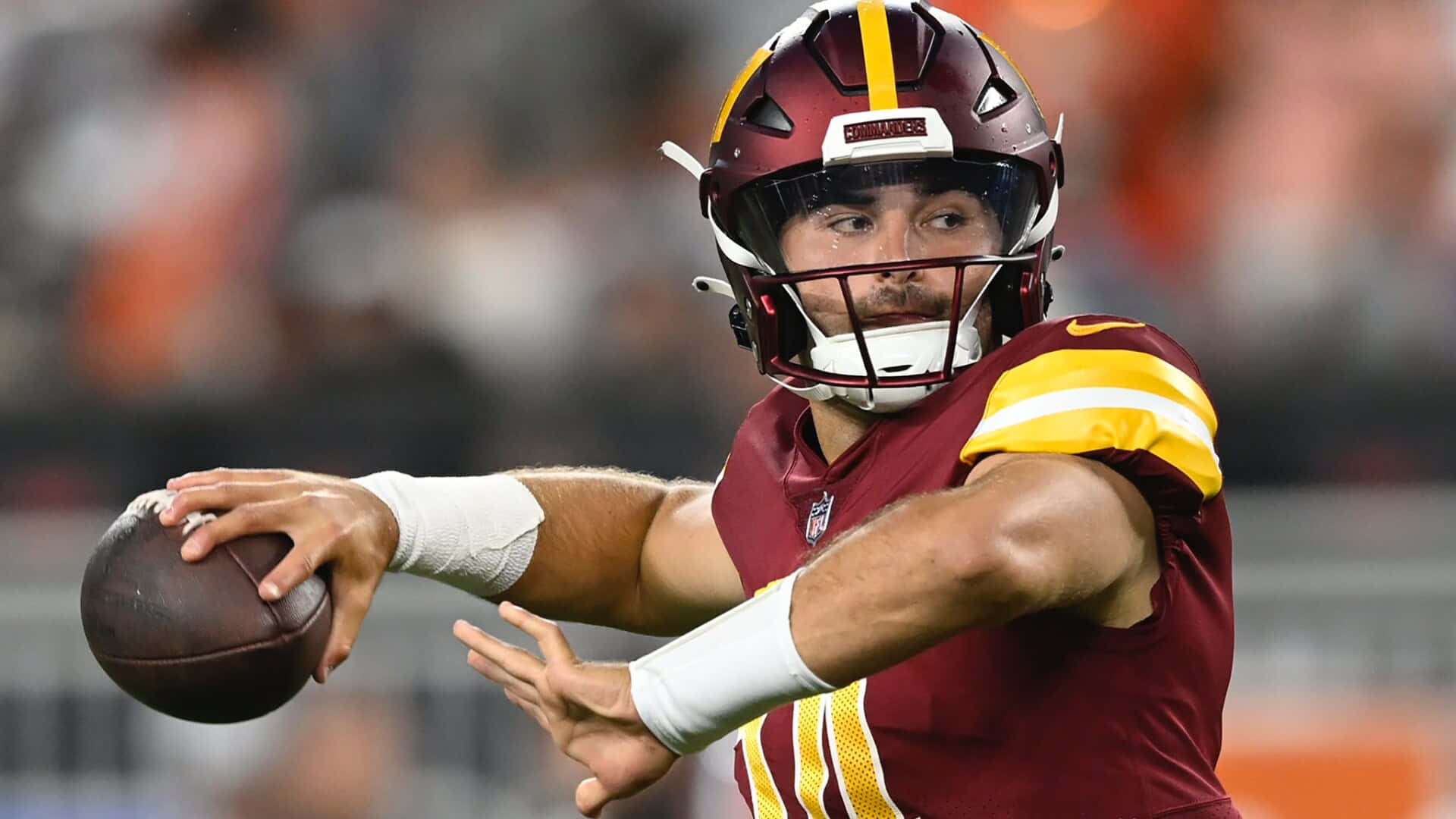  壁紙 — 一位專注的NFL四分衛身穿棗紅色和金色球衣，在球場燈光下準備擲出橄欖球。