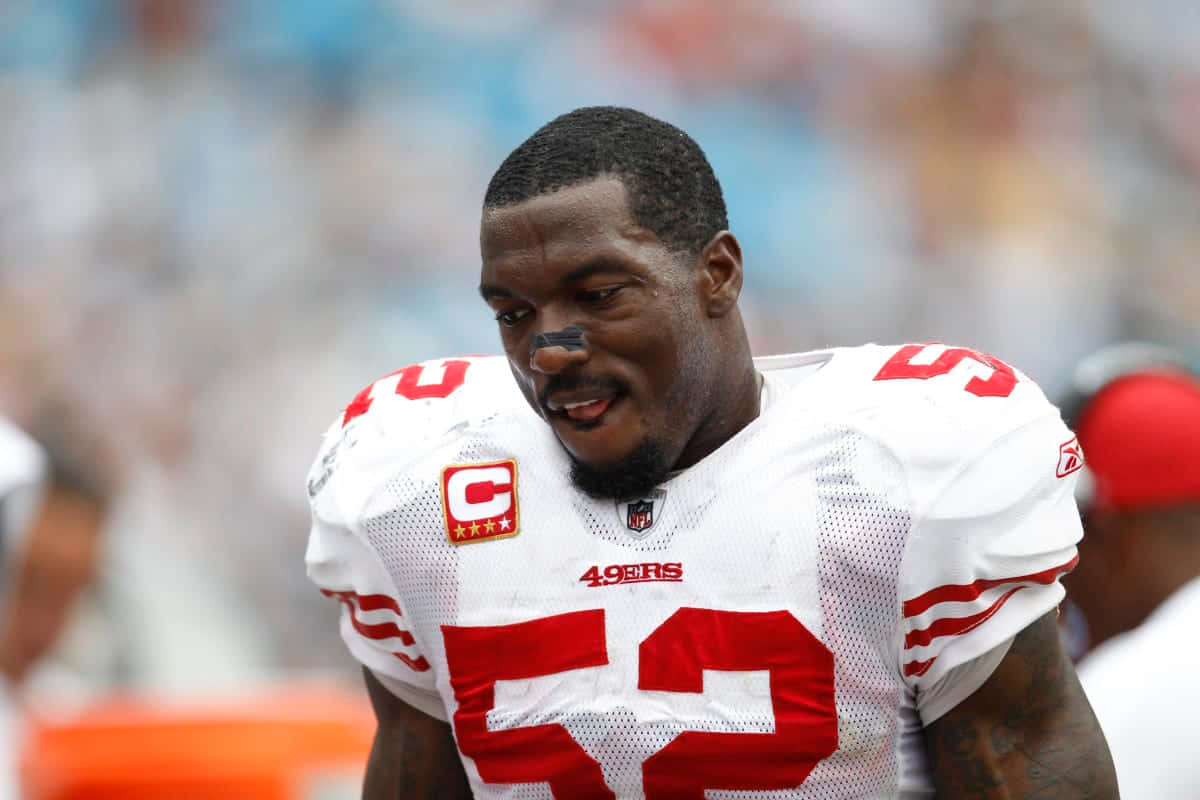  壁紙 — 一位專注的NFL線衛身穿白色和紅色的49ers球衣，號碼52，在日間比賽中低頭。